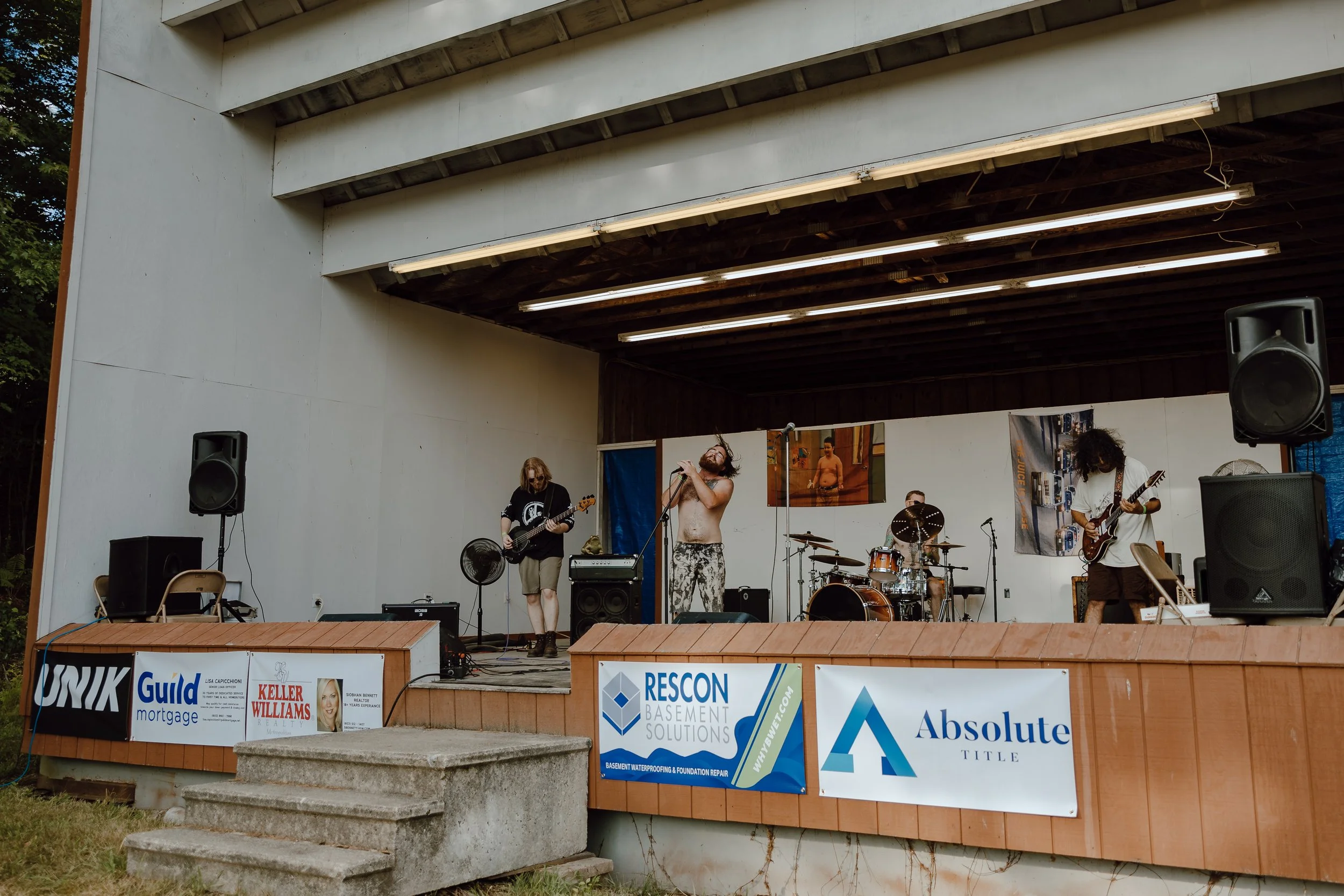 A band performing live on an outdoor stage with four members: one playing guitar, one singing into a microphone, one playing drums, and one playing bass guitar. The stage has banners with various advertisements on the front.