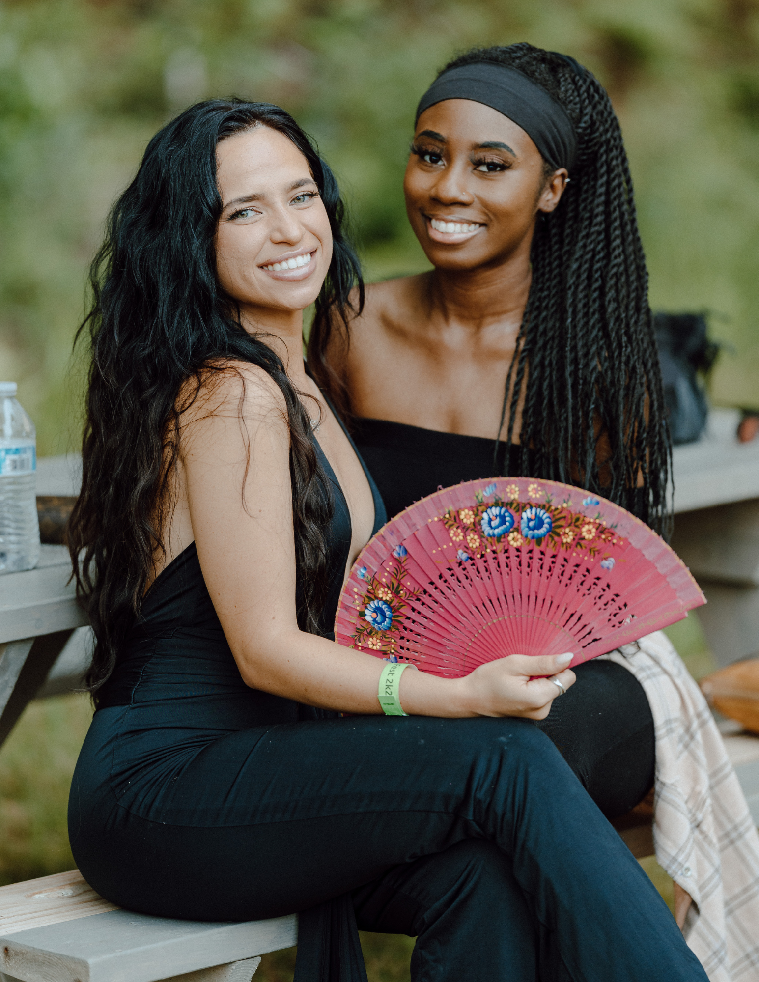 Two women sitting on a park bench, smiling, with one holding a pink decorative fan.