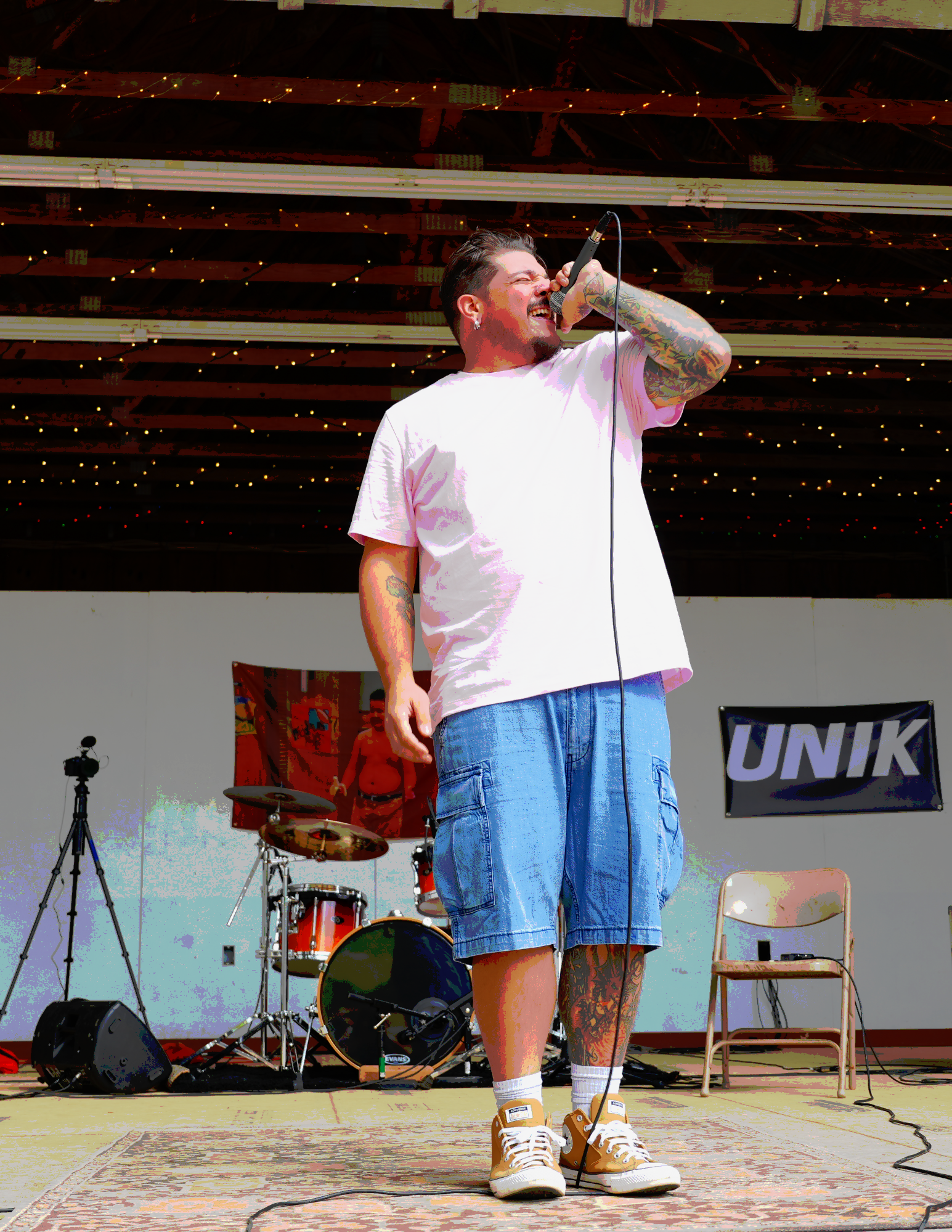 A male singer passionately performs on stage, holding a microphone with his right hand, wearing a white T-shirt, denim shorts, and sneakers, with tattoos on his left arm and leg. There is musical equipment, including drum set and microphone stand, in the background.