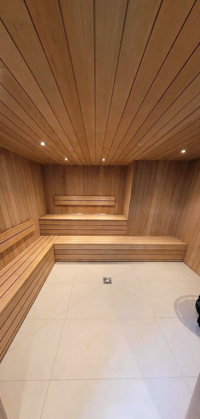 A room with wood-paneled walls and ceiling, featuring built-in wooden benches around the perimeter and a tiled floor with a drain in the center.