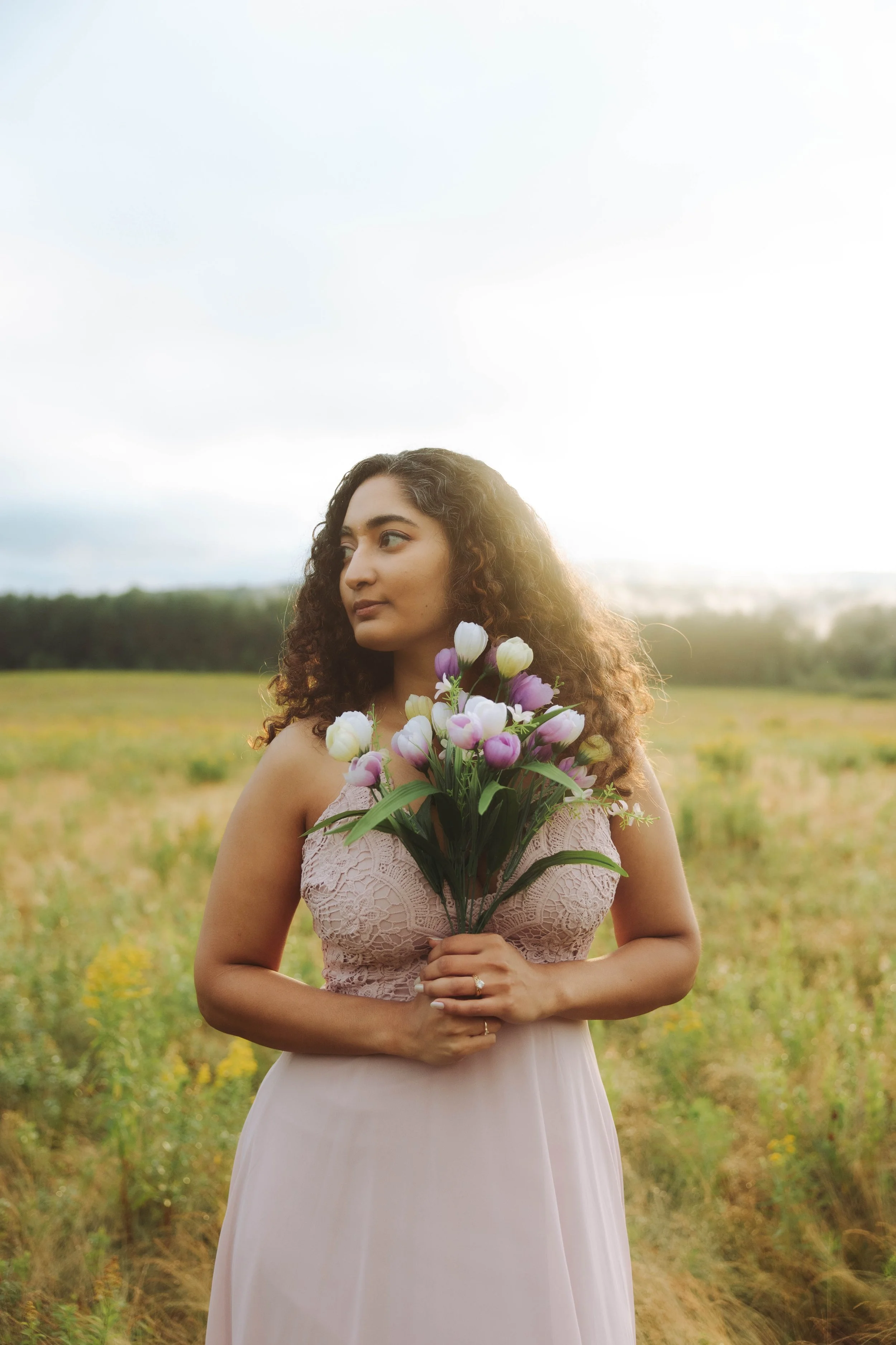 A woman standing outdoors in a field, holding a bouquet of purple and white flowers, wearing a pink lace dress, with a soft sunlight glow behind her.