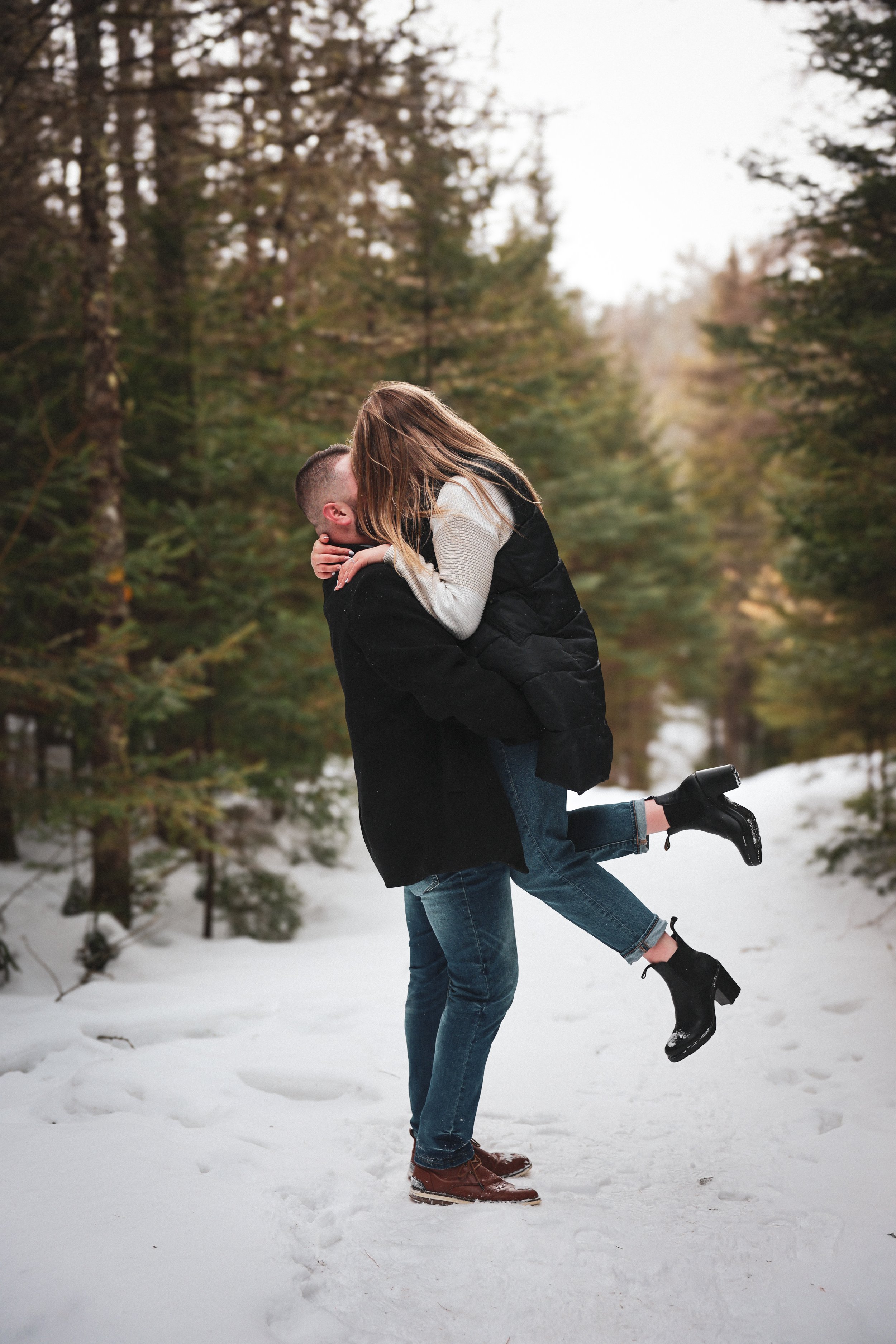 A couple is outdoors in a snowy forest, with the man lifting the woman off the ground while kissing.