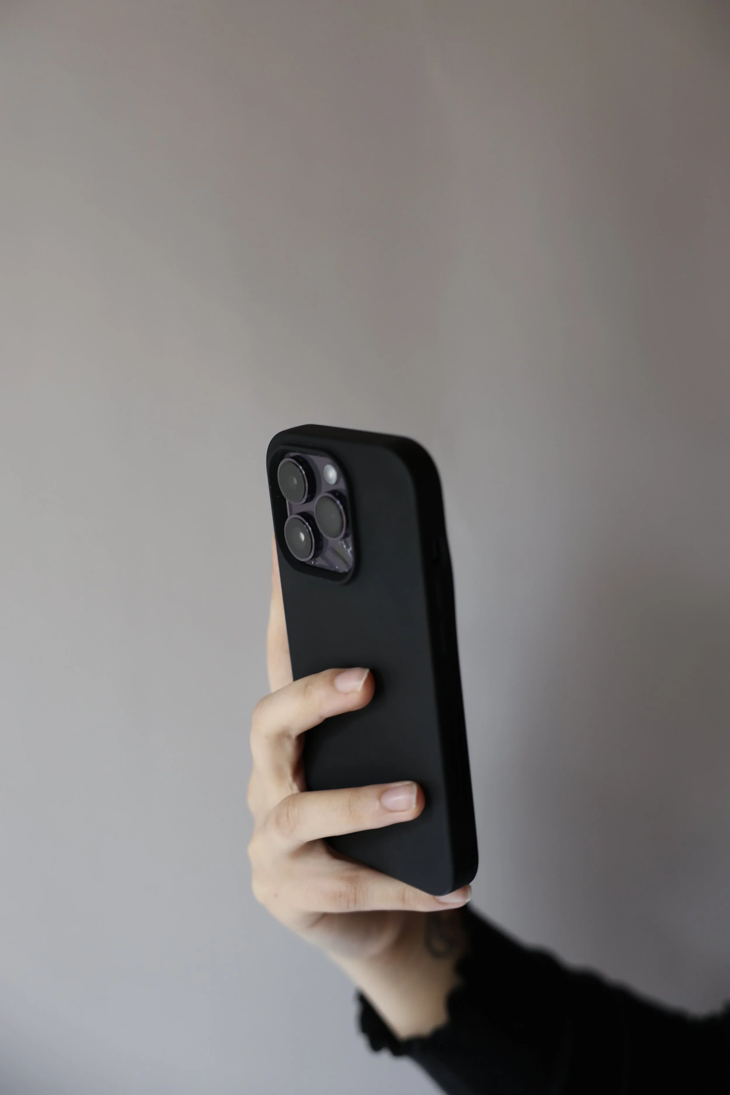 A person holding a black smartphone with a triple camera setup, against a plain gray background.