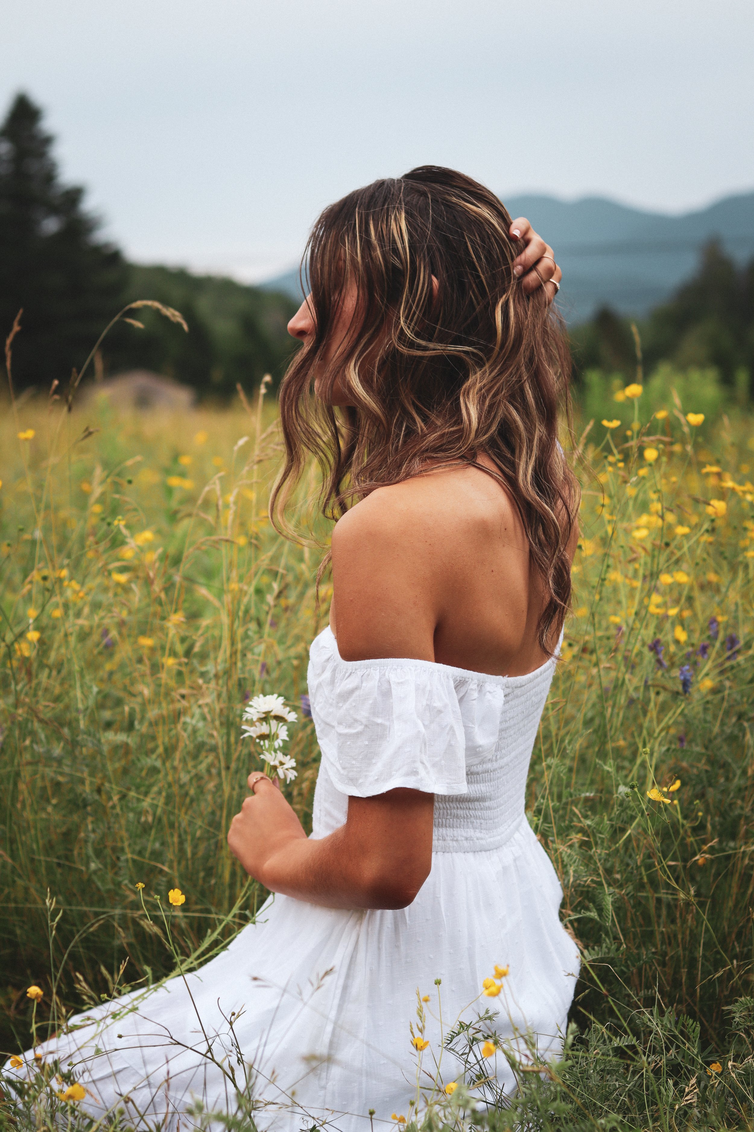 A woman in a white off-shoulder dress sitting in a meadow with yellow flowers, holding a small bunch of white daisies, with mountains and trees in the background.