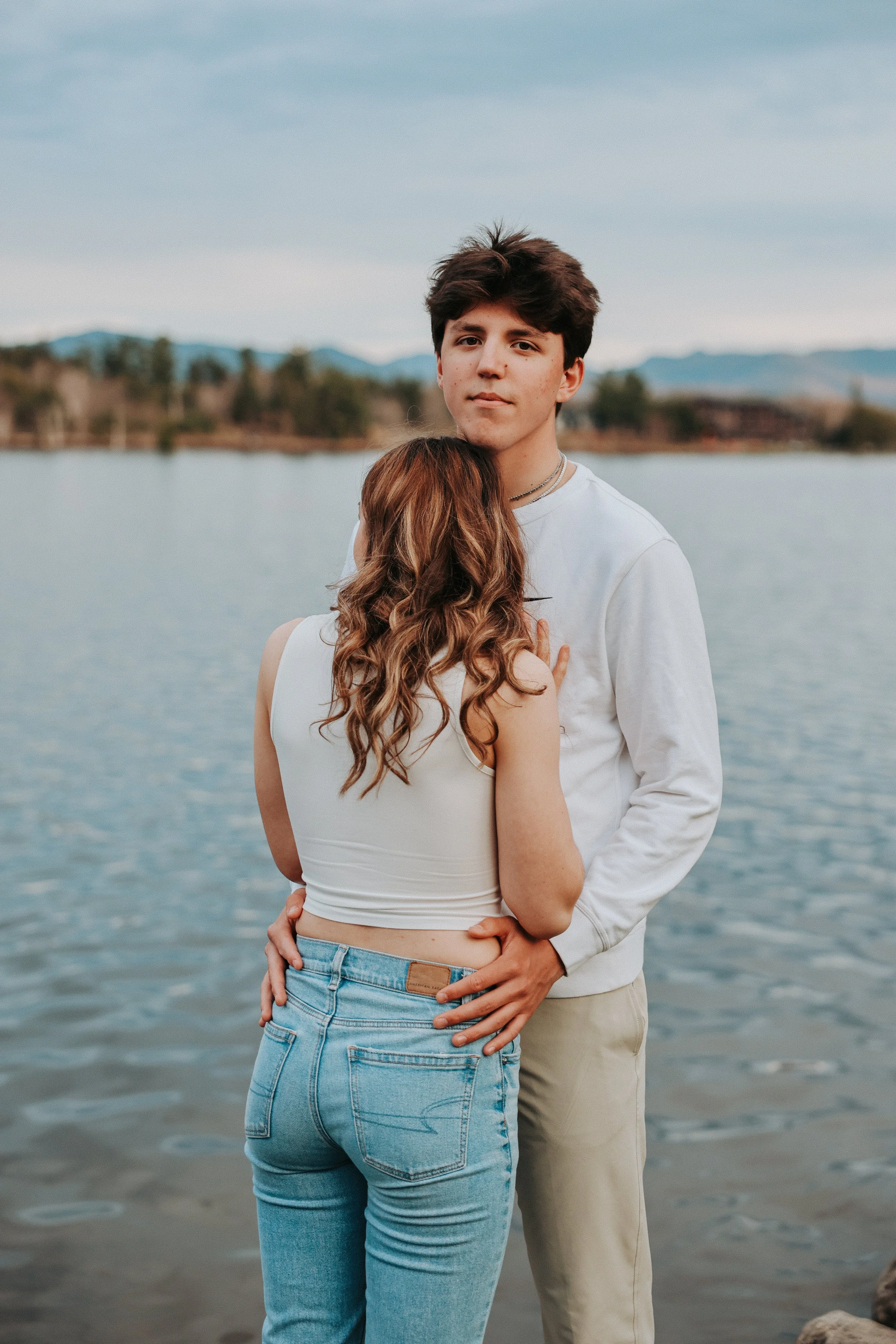 A young man and woman standing by a lake, with trees and mountains in the background. The woman is hugging the man from in front, and only her back is visible. The man has a neutral expression and is looking at the camera.