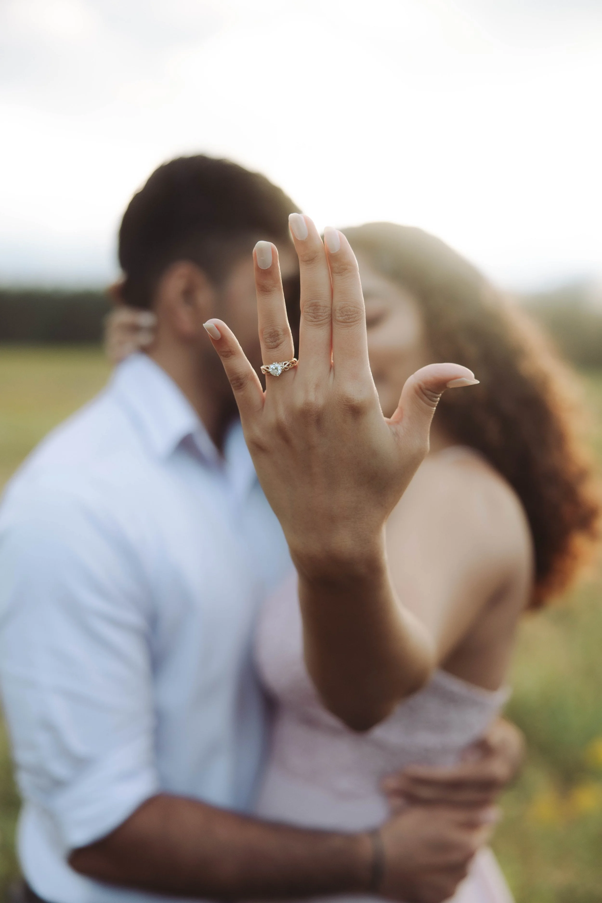 A woman showing off an engagement ring on her finger as a couple kisses in the background outdoors.
