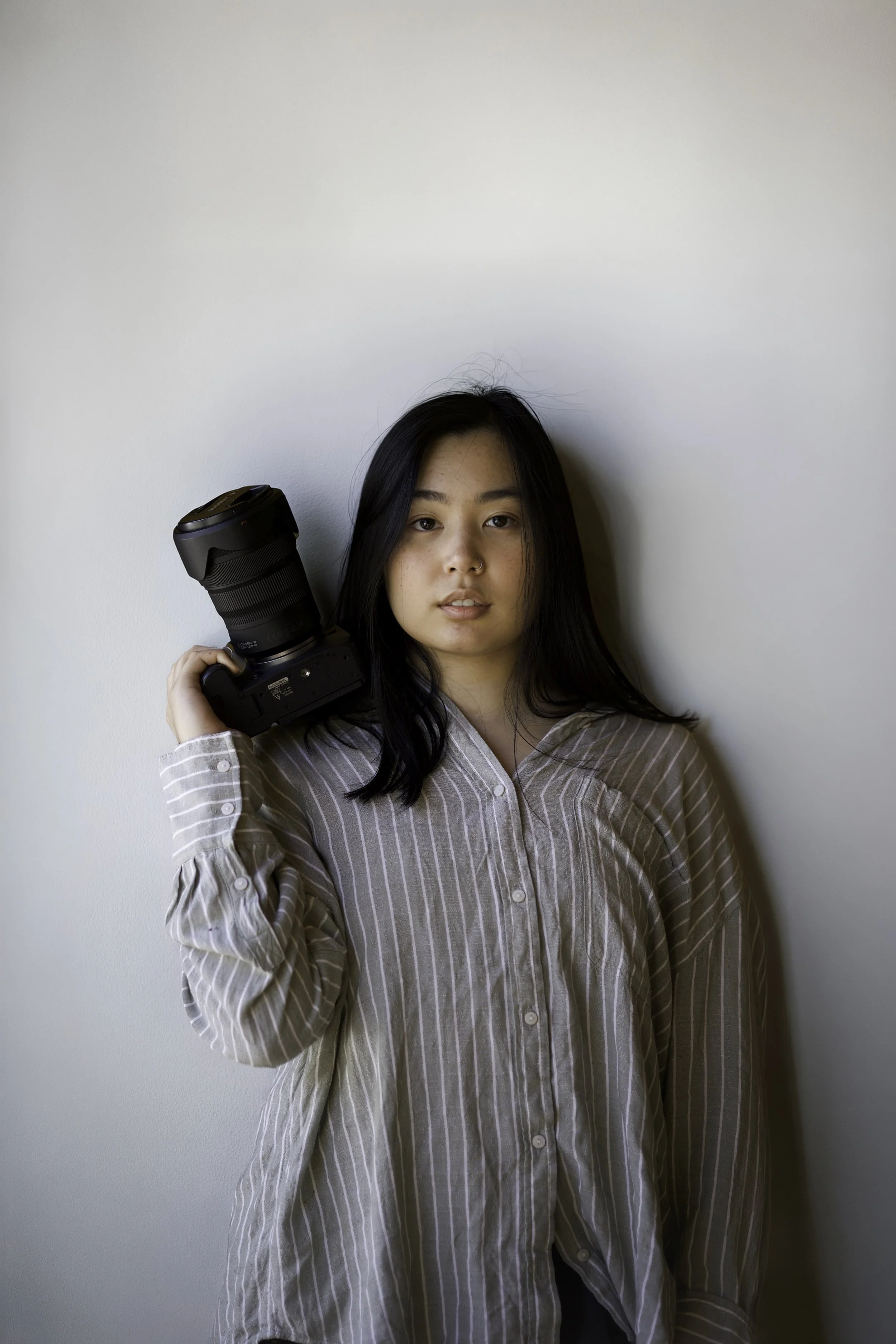 Young woman with black hair wearing a beige striped shirt holding a camera with a large lens against a plain white wall.