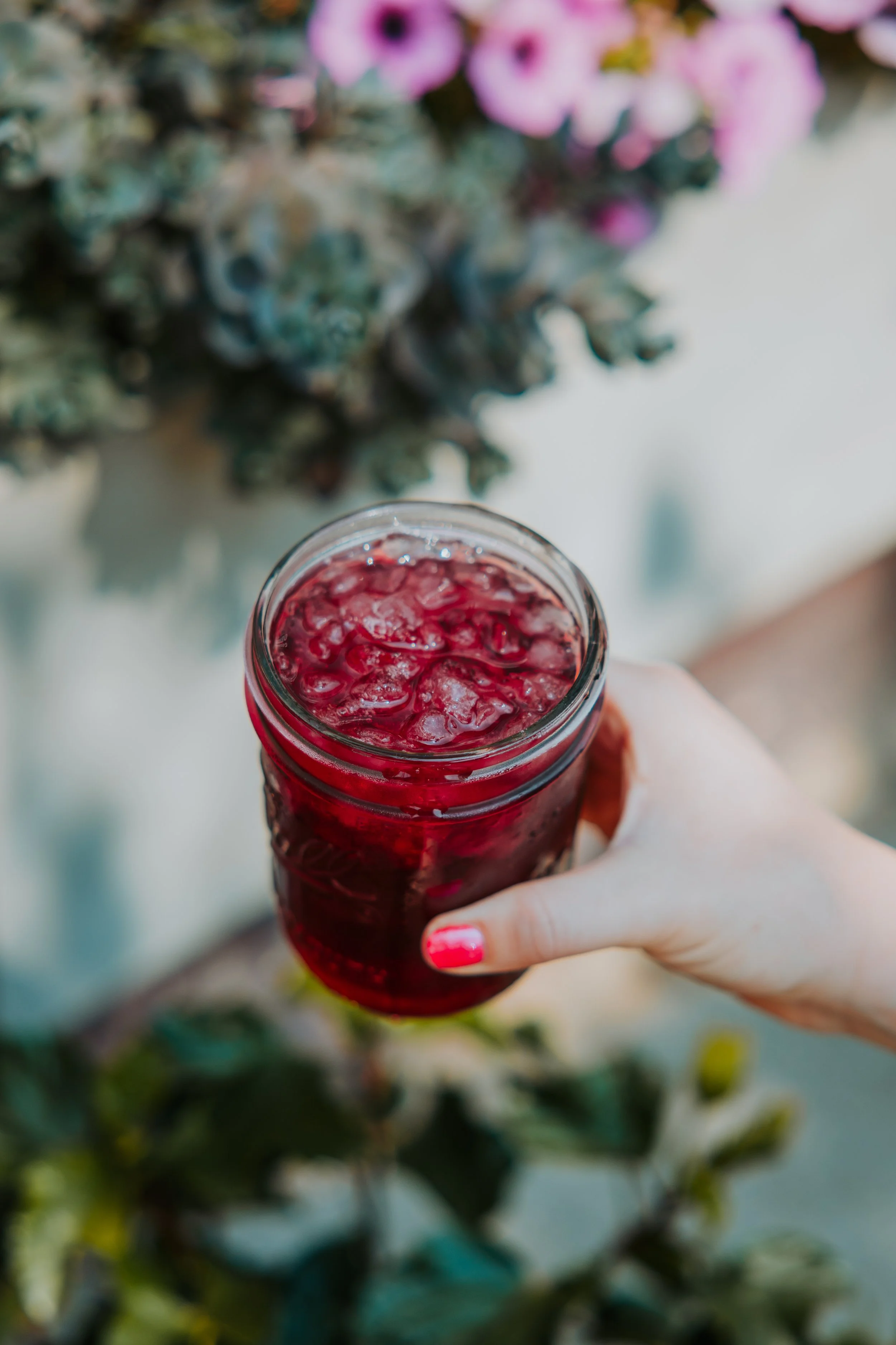 A person's hand with pink painted nails holding a glass jar filled with red, ice-filled beverage, with pink flowers and greenery in the blurred background.
