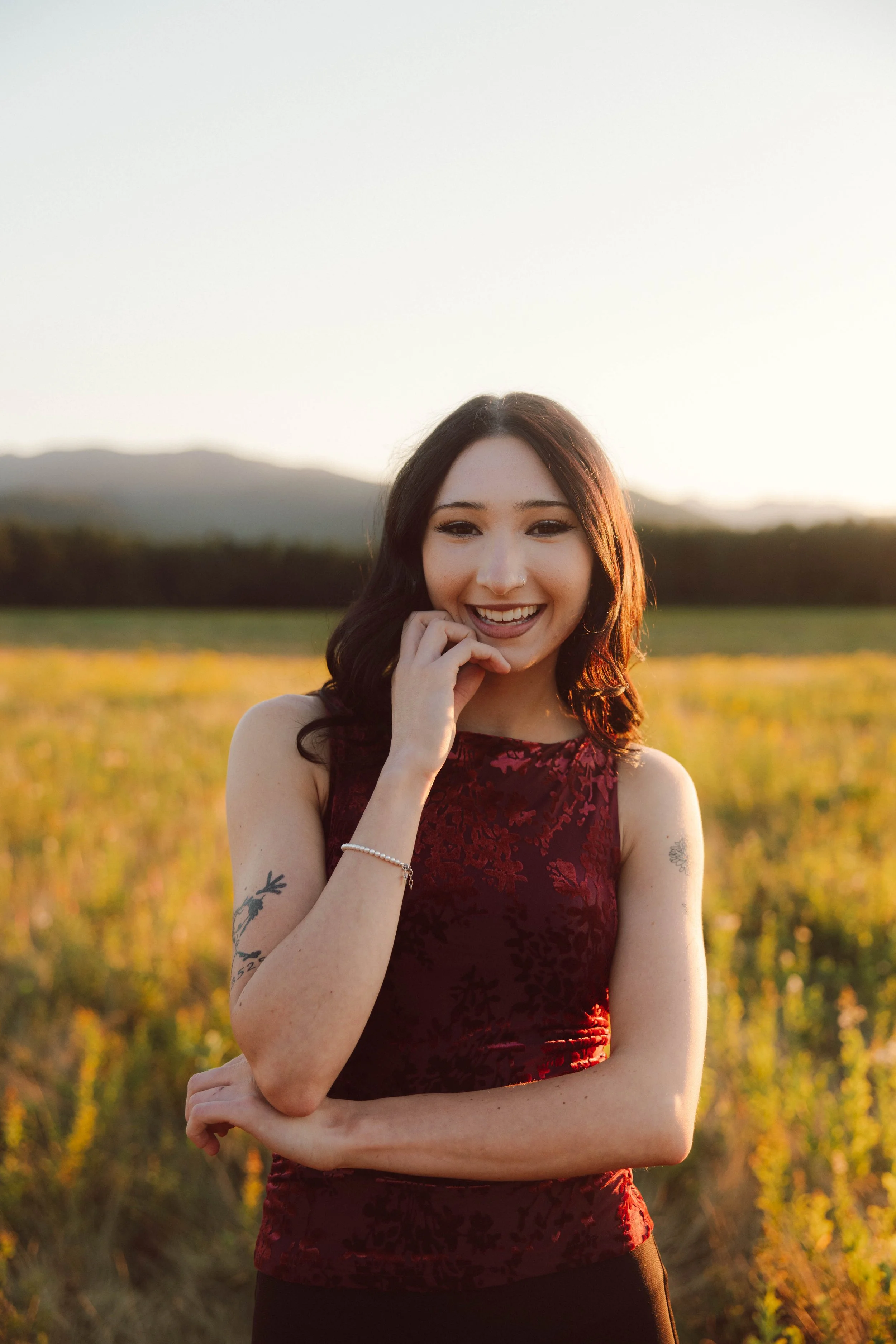 A woman with dark hair and tattoos on her arms, wearing a sleeveless red dress, smiling outdoors during sunset in a field.