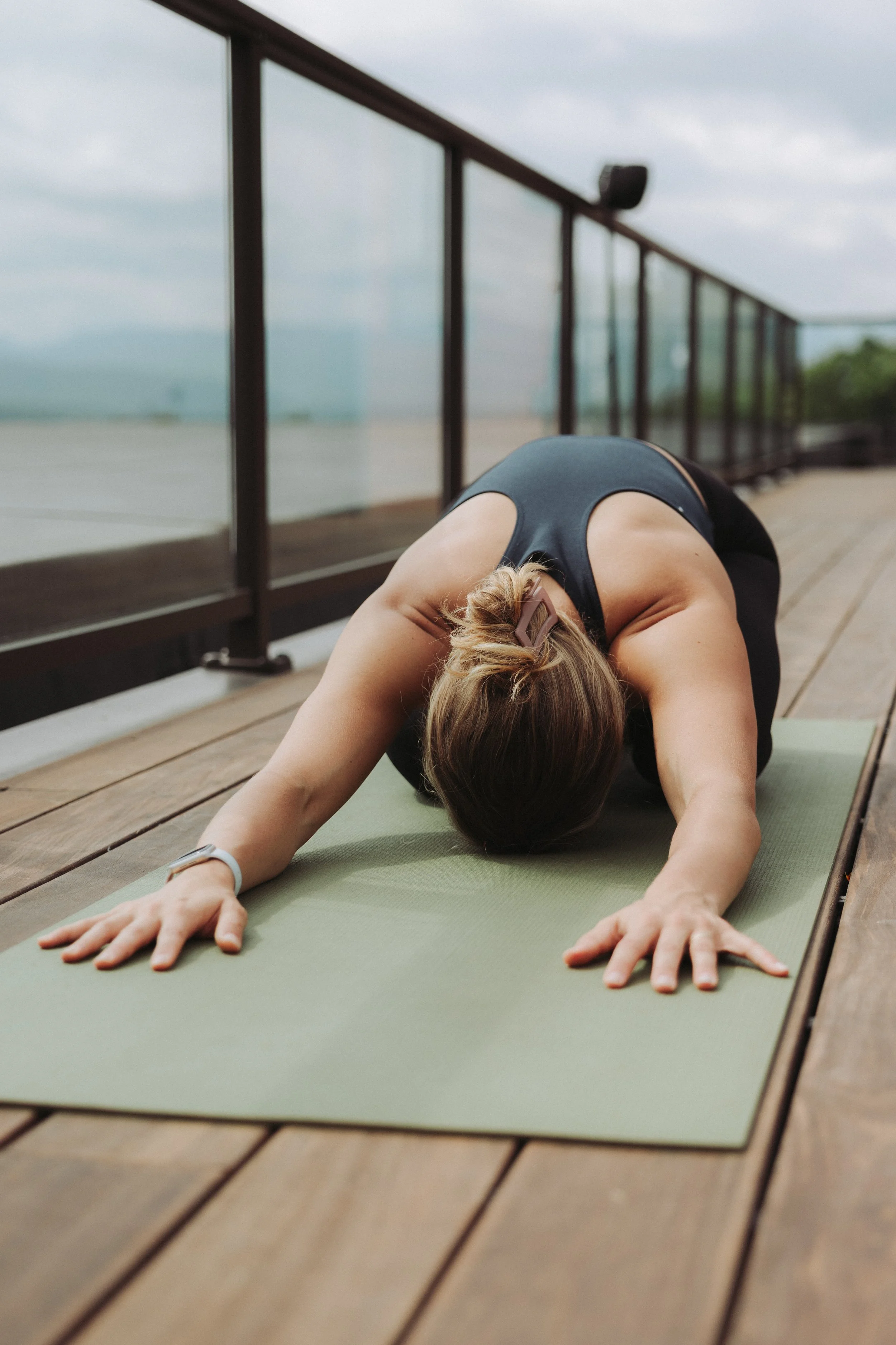 A woman practicing yoga outdoors on a green mat on a wooden deck, stretching in child's pose with her arms extended forward and head on the mat, surrounded by cloudy skies and greenery in the background.
