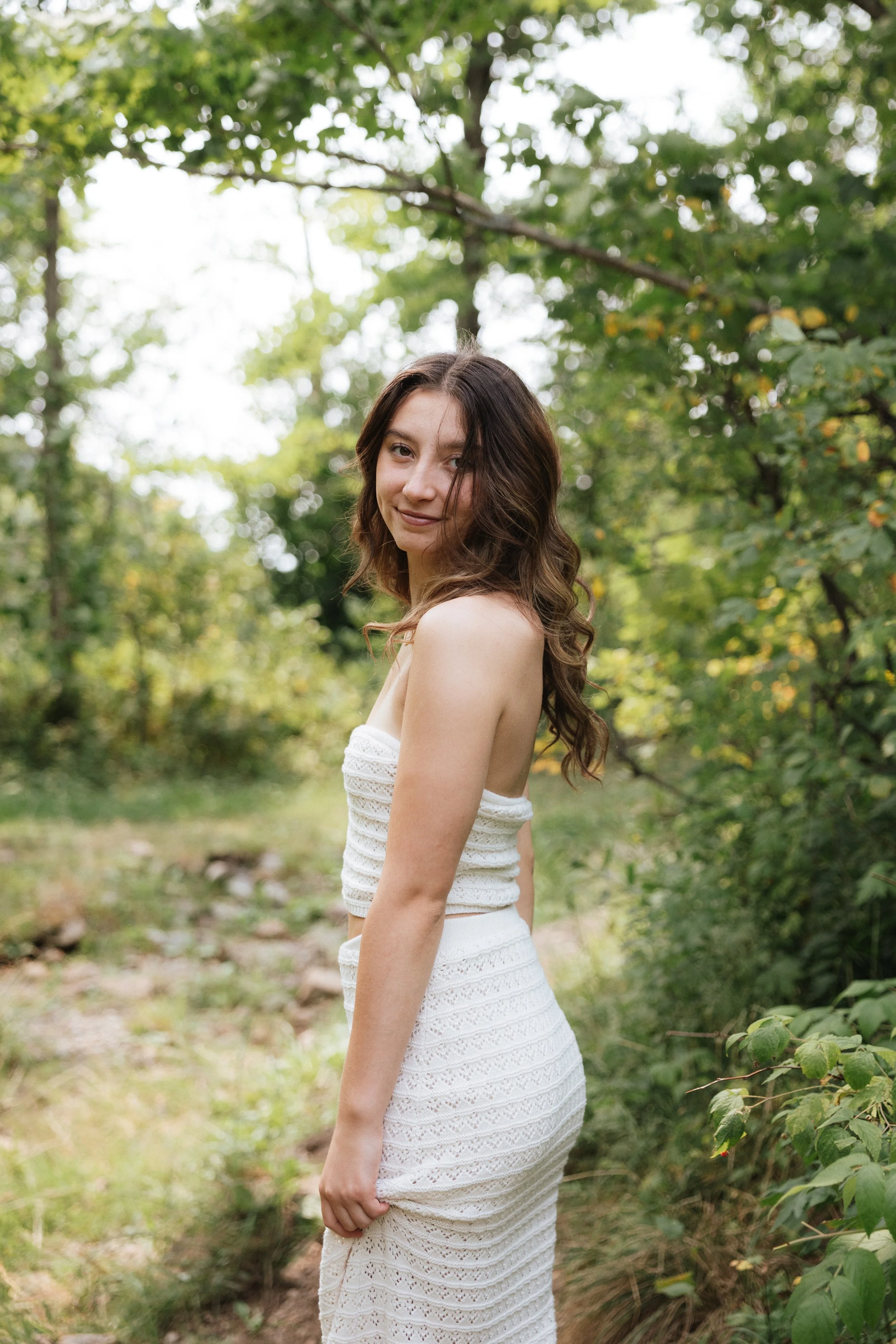 A young woman with brown hair wearing a white strapless dress stands outdoors on a forest trail, smiling at the camera, surrounded by green trees and foliage.