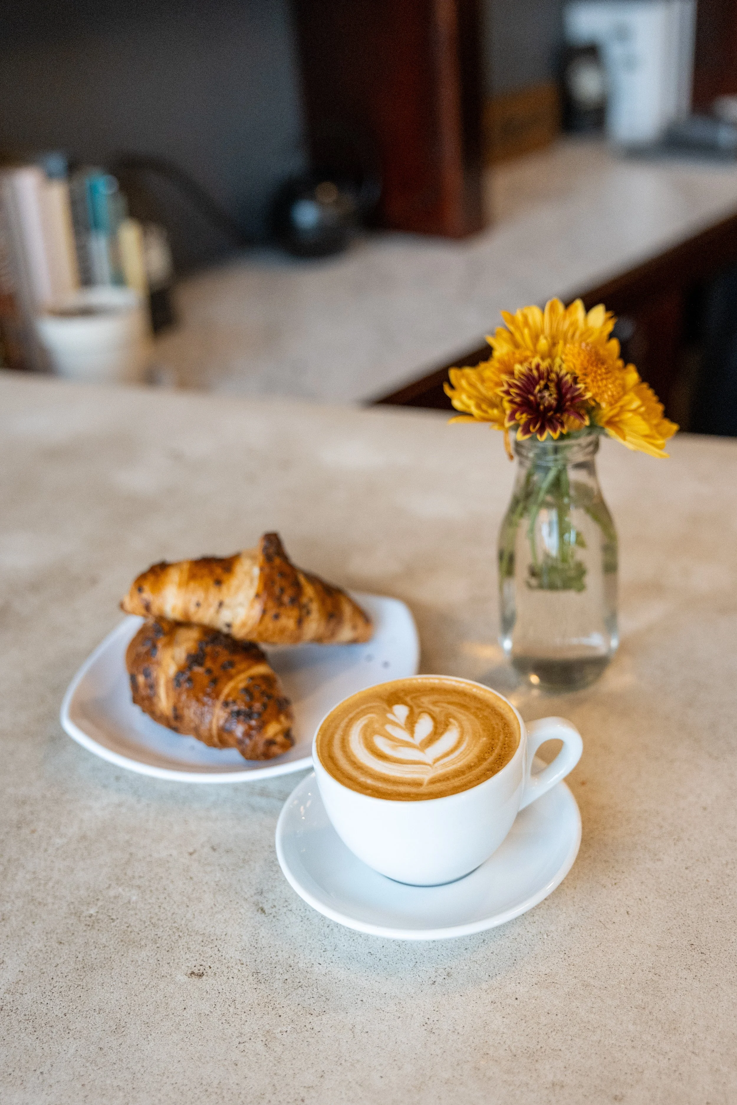 A cup of latte with latte art on top, two croissants on a white plate, and a small glass vase with yellow and maroon flowers on a beige countertop.