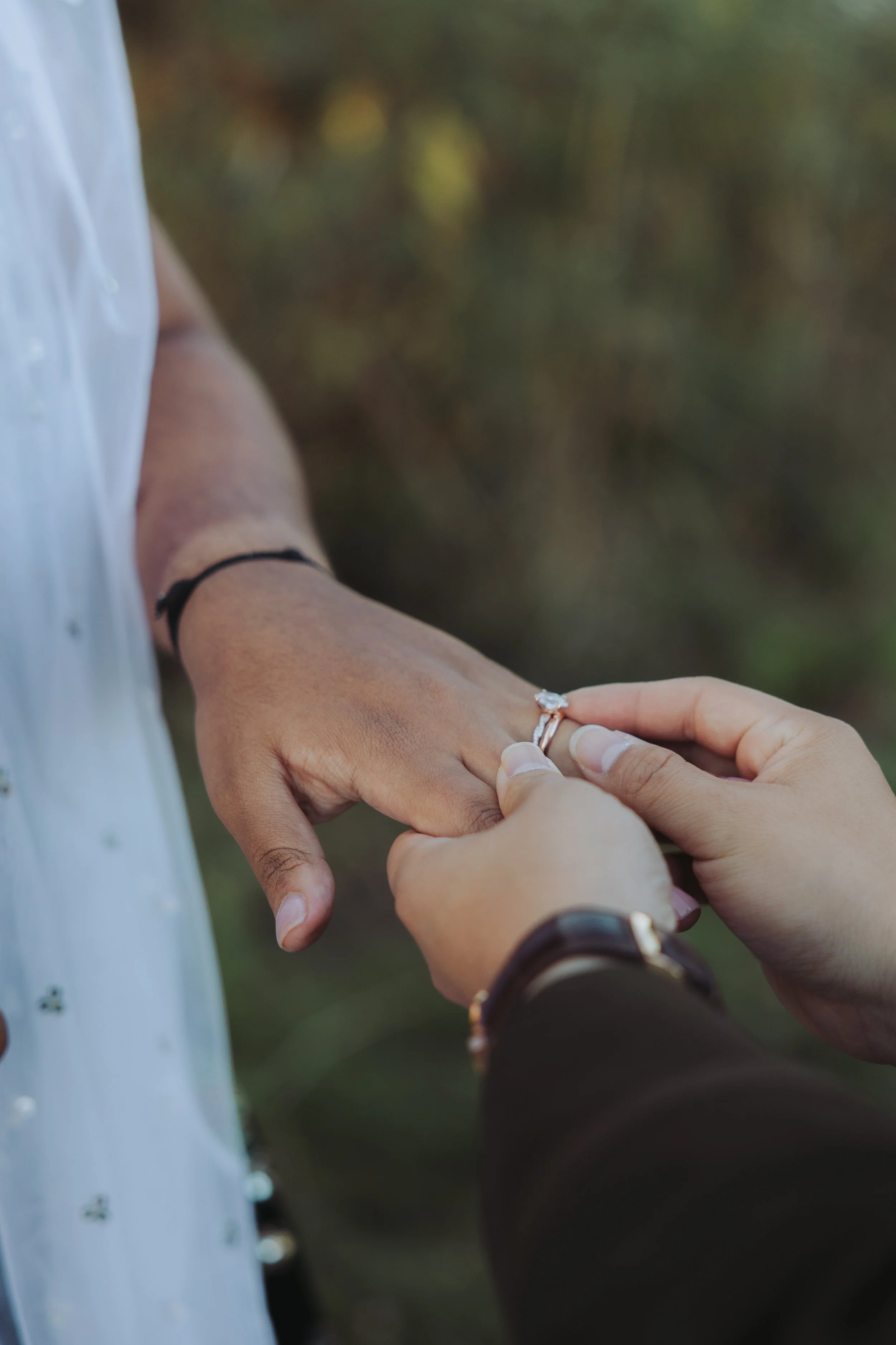 Person placing an engagement ring on another person's finger outdoors.