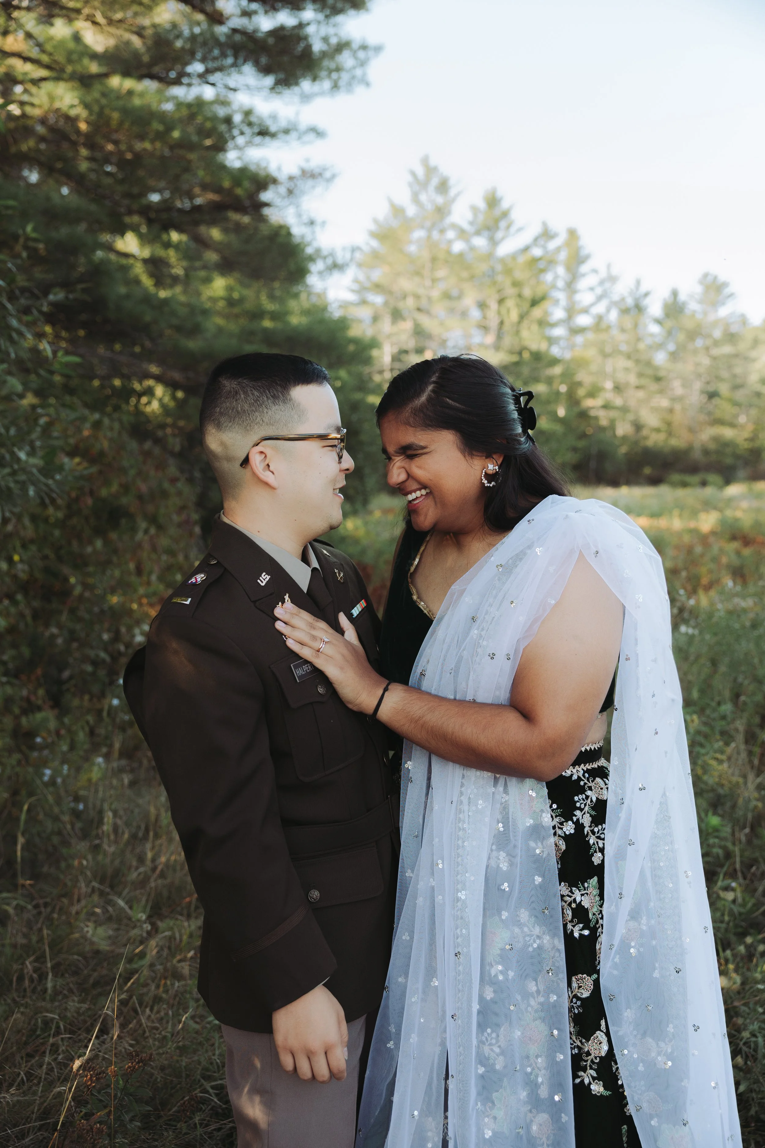 A person in a military uniform and a woman in a white and black dress share a joyful moment outdoors, smiling and touching foreheads.