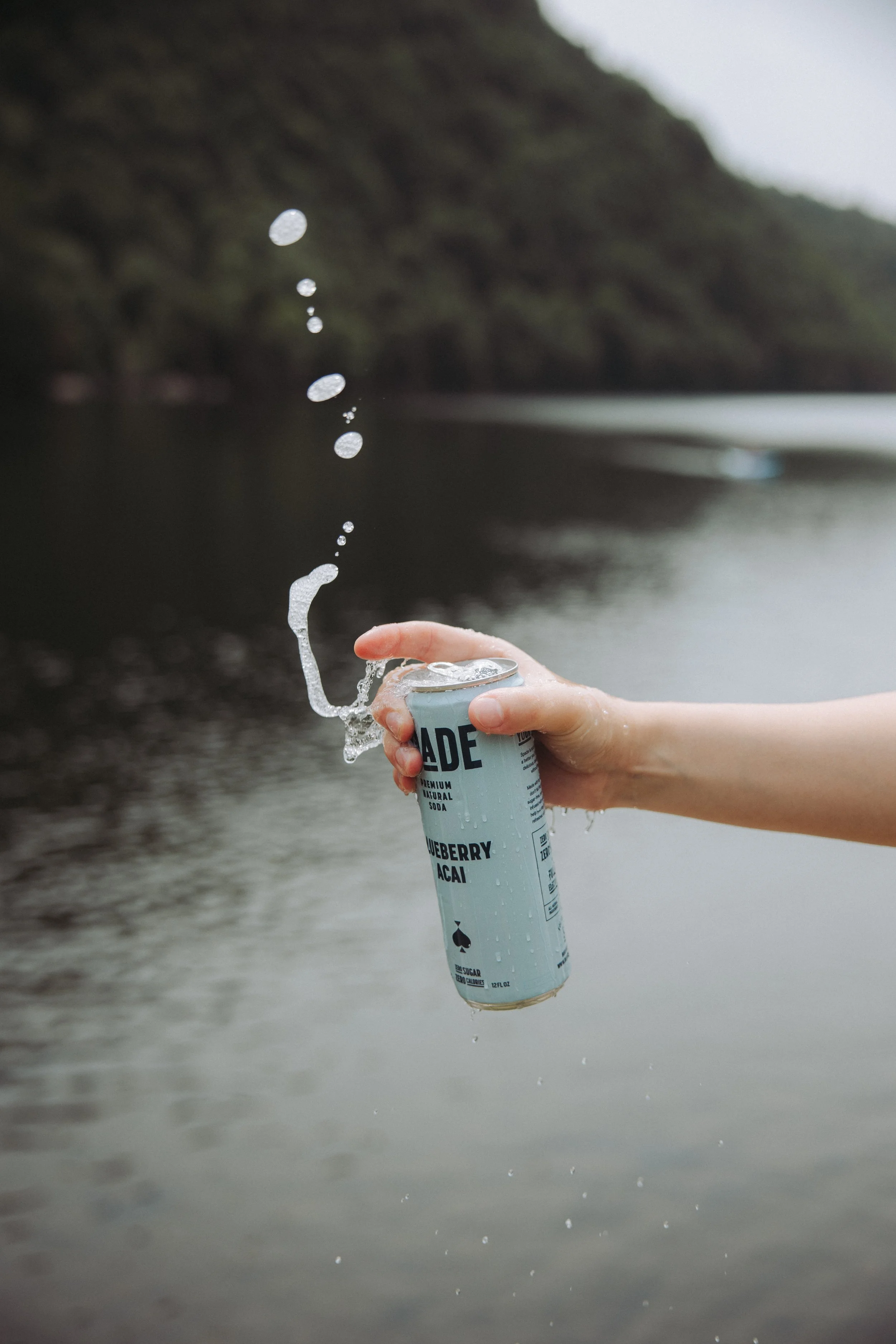 A person holding a can of blueberry acai flavored sparkling water over a body of water, with water splashing out as the can is opened.