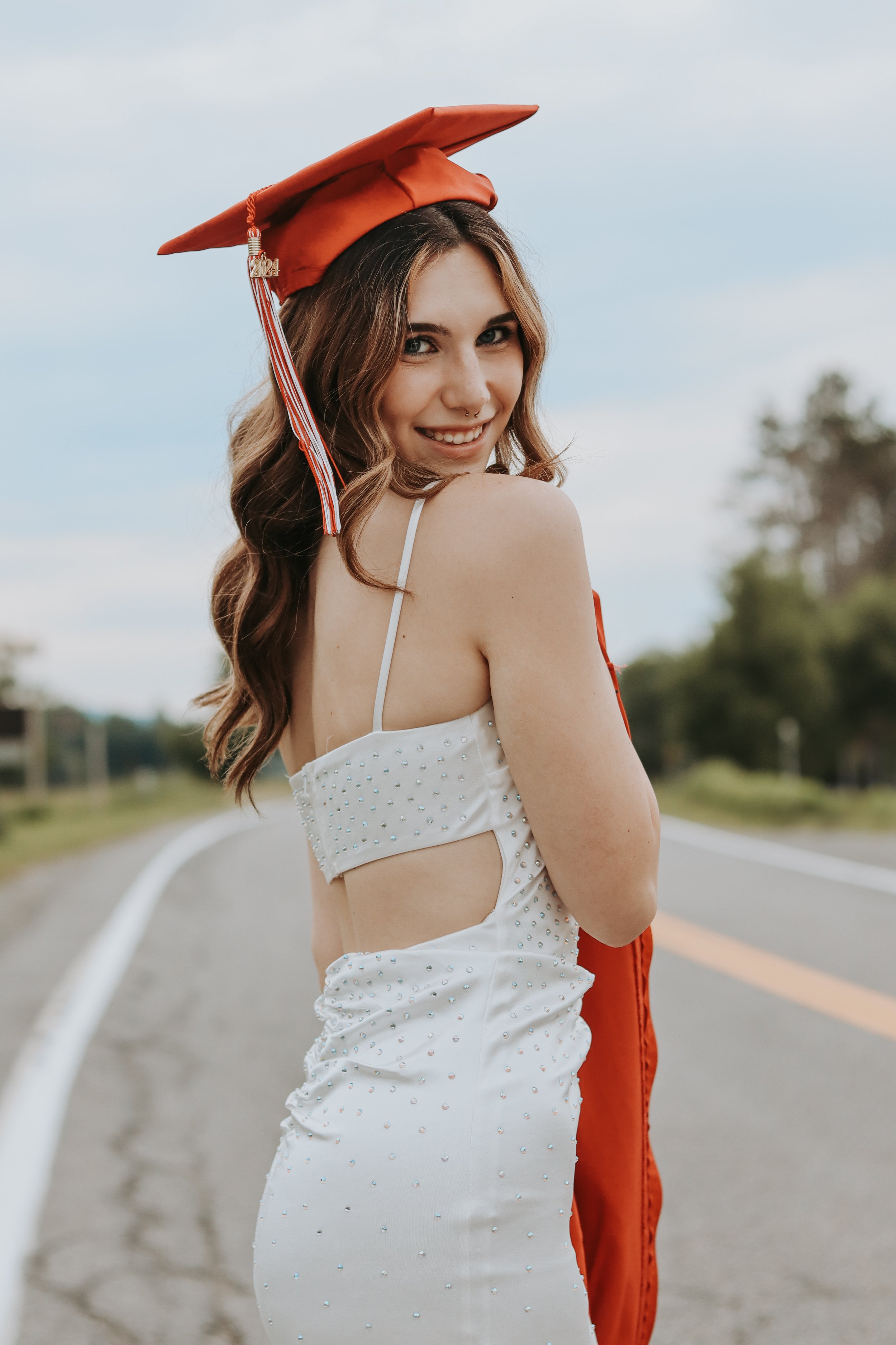 A young woman smiling in a white dress with studded details, wearing an orange graduation cap, standing on an empty rural road with green trees in the background.