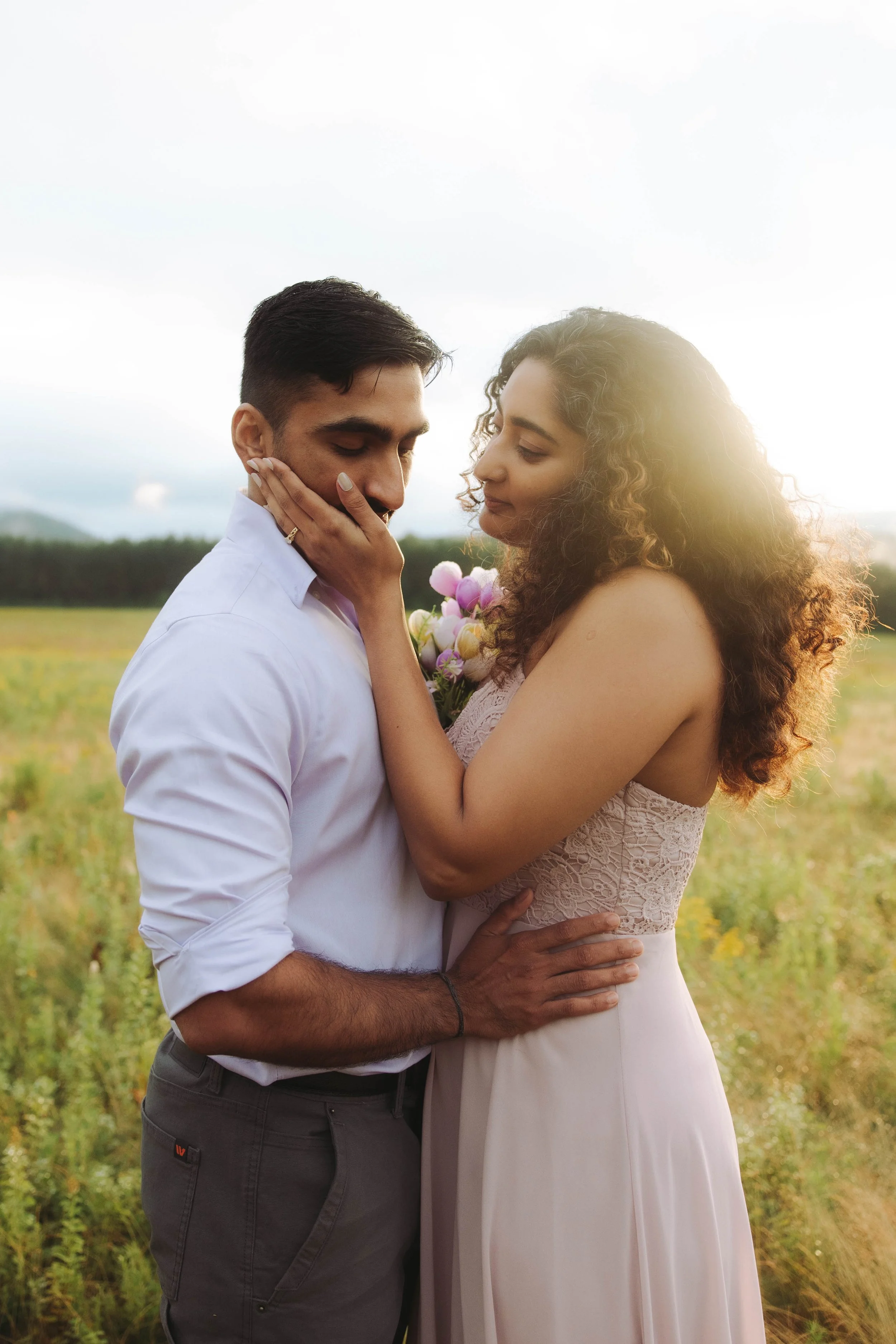 A man and woman embracing in a field during sunset, with the woman holding flowers and gently touching the man's face.