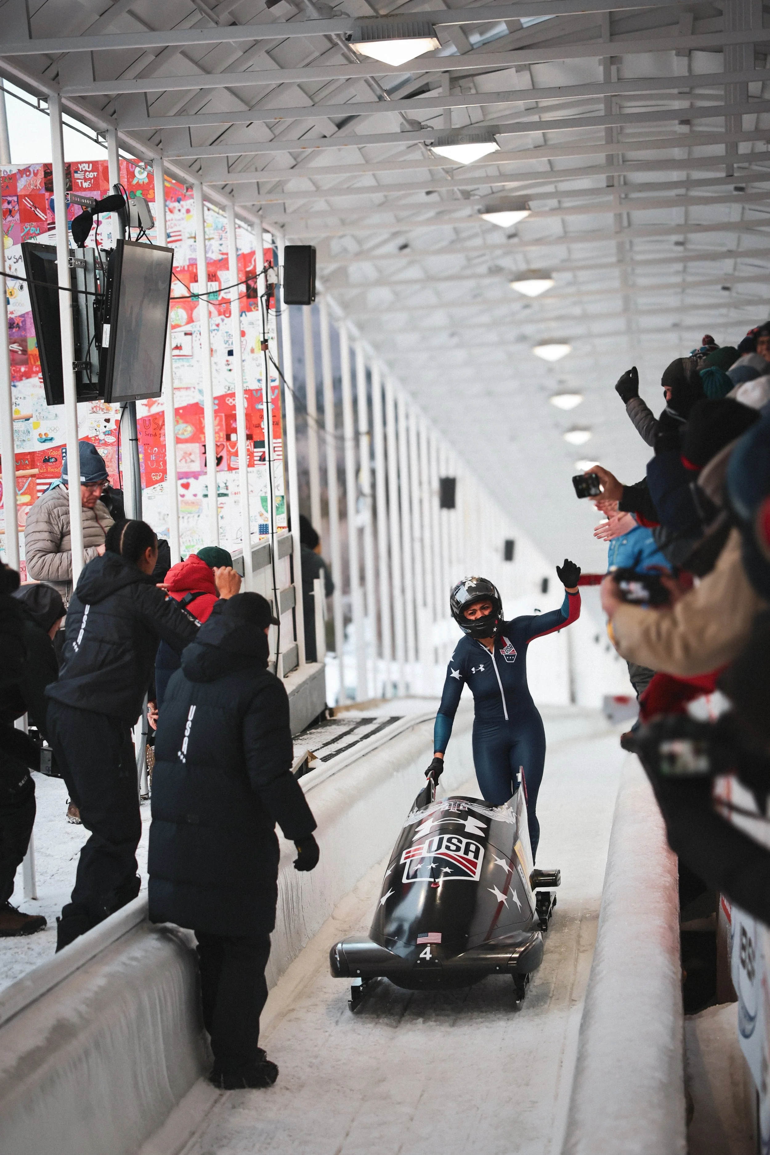 A female bobsled athlete, wearing a US team uniform and protective helmet, waves to spectators after racing down the track in a bobsled with US and American flag decals. Spectators in winter clothing cheer and take photos along the track at a winter 