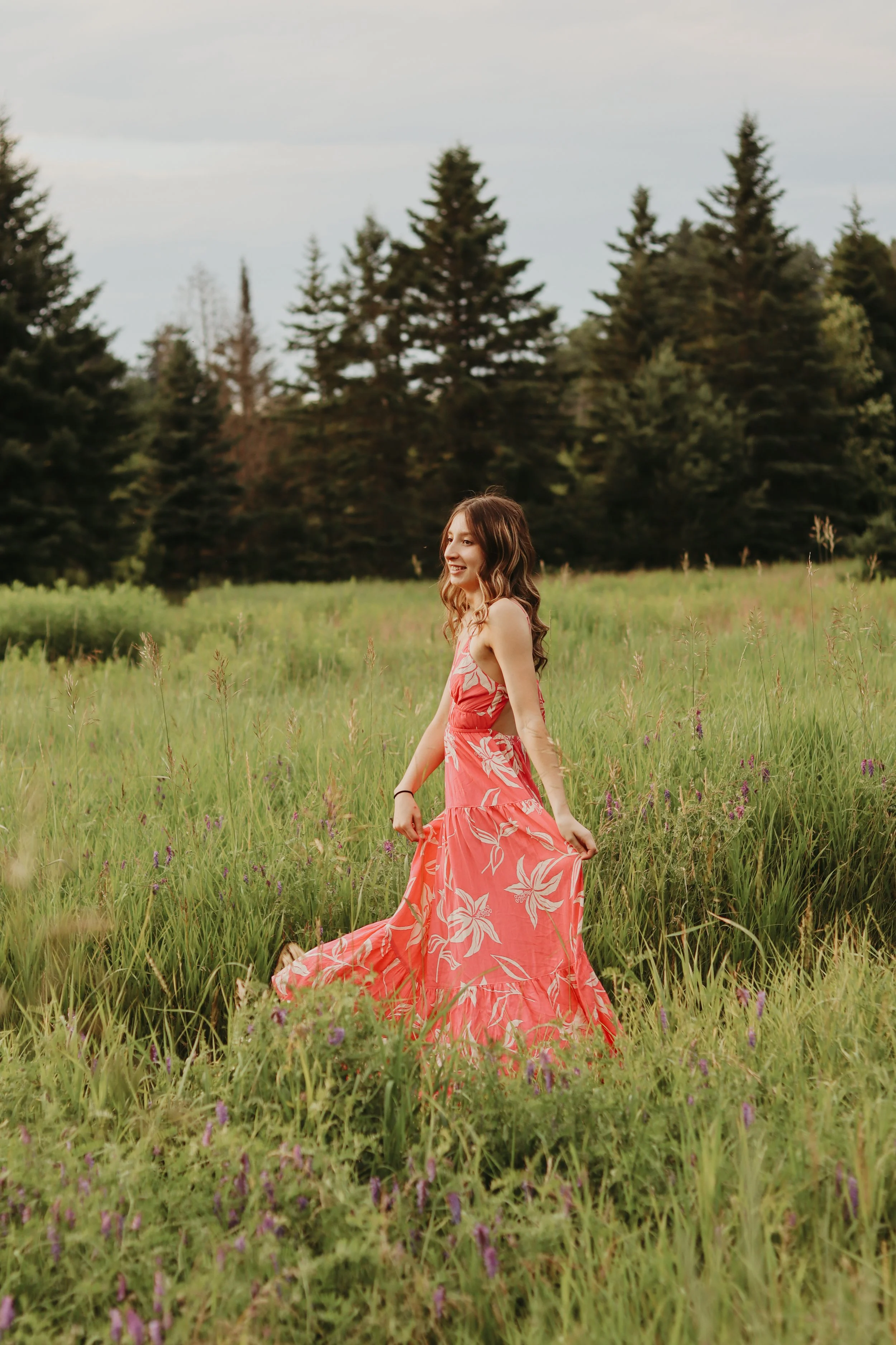 A woman in a pink floral dress walking through a grassy field with purple flowers, against a backdrop of trees and a cloudy sky.
