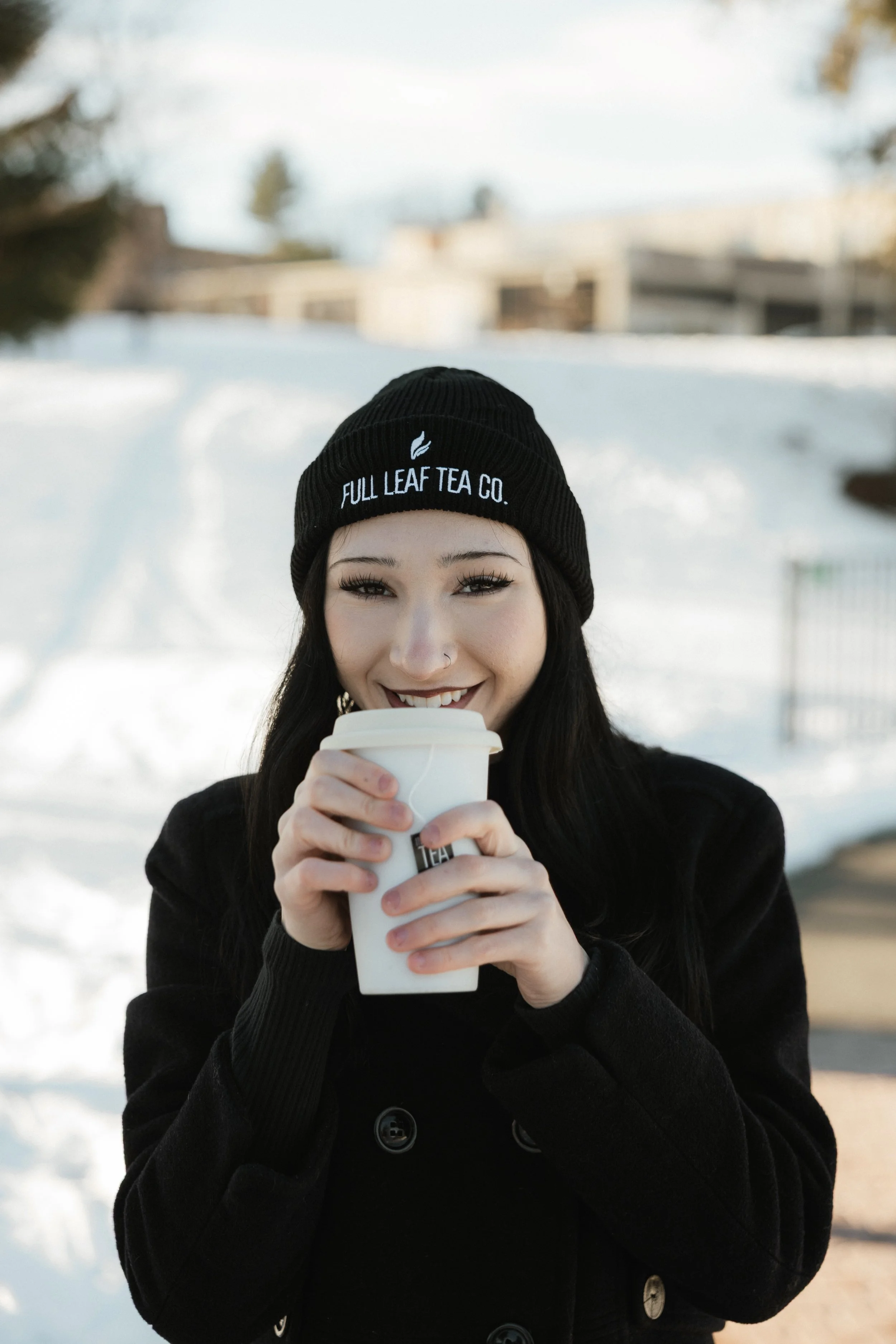 A woman in a black beanie and coat enjoying a hot drink outdoors in a snowy setting.