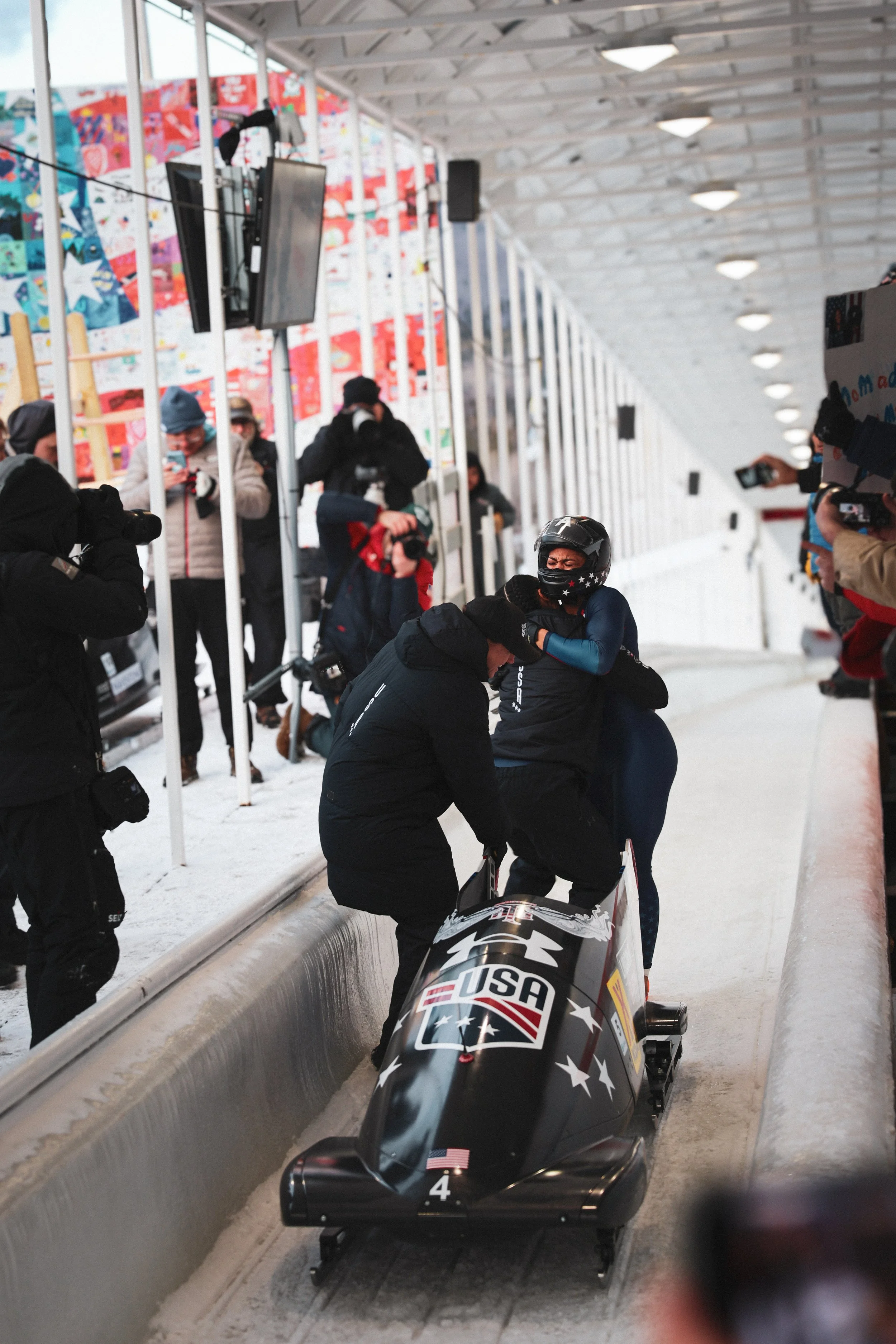 A bobsled team celebrating a victory inside a snowy indoor track, with spectators and photographers capturing the moment.