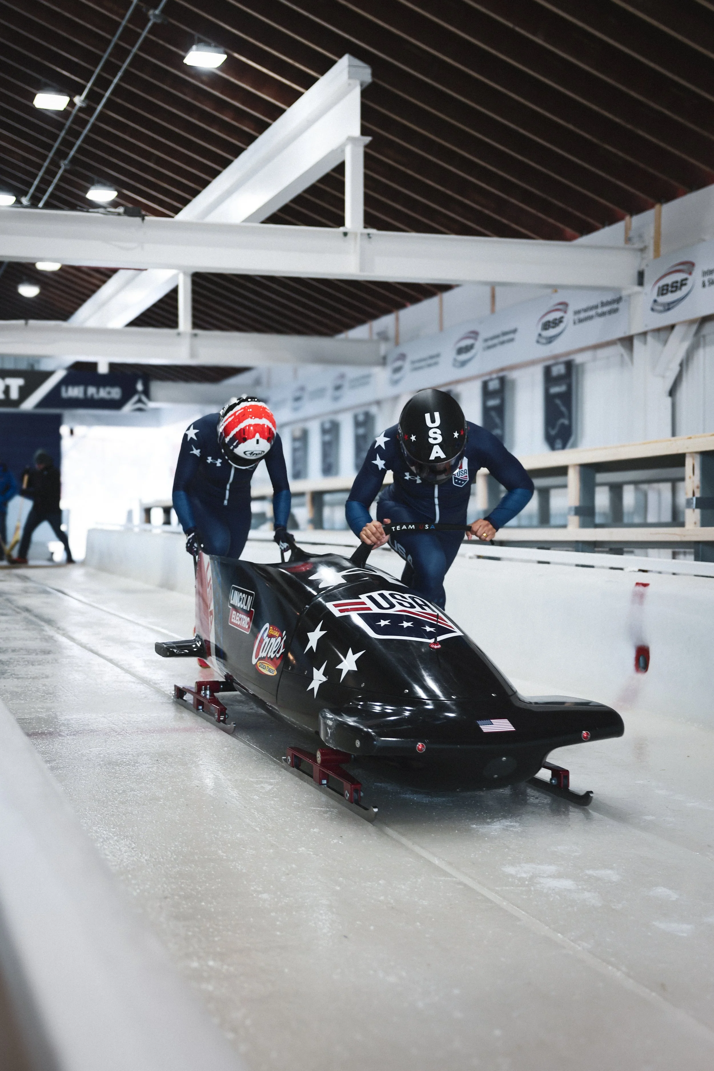 USAsled bobsled team members pushing their black and red bobsled down an indoor ice track, wearing blue suits with stars and helmets with 'USA' and stars, inside a sports facility.