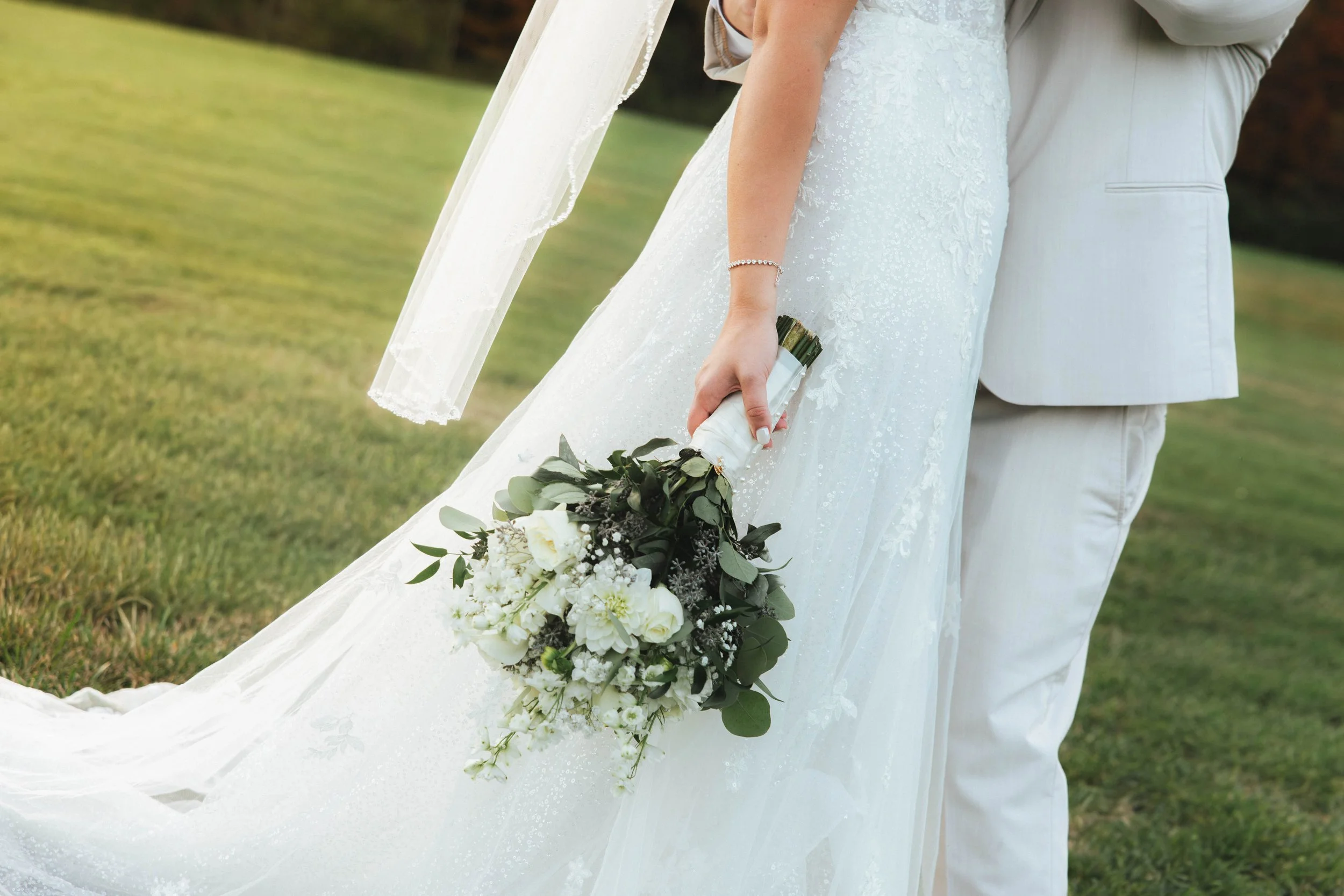 Bride holding a bouquet of white flowers and greenery, dressed in a white wedding gown, beside a groom in a light-colored suit, on a grassy lawn.