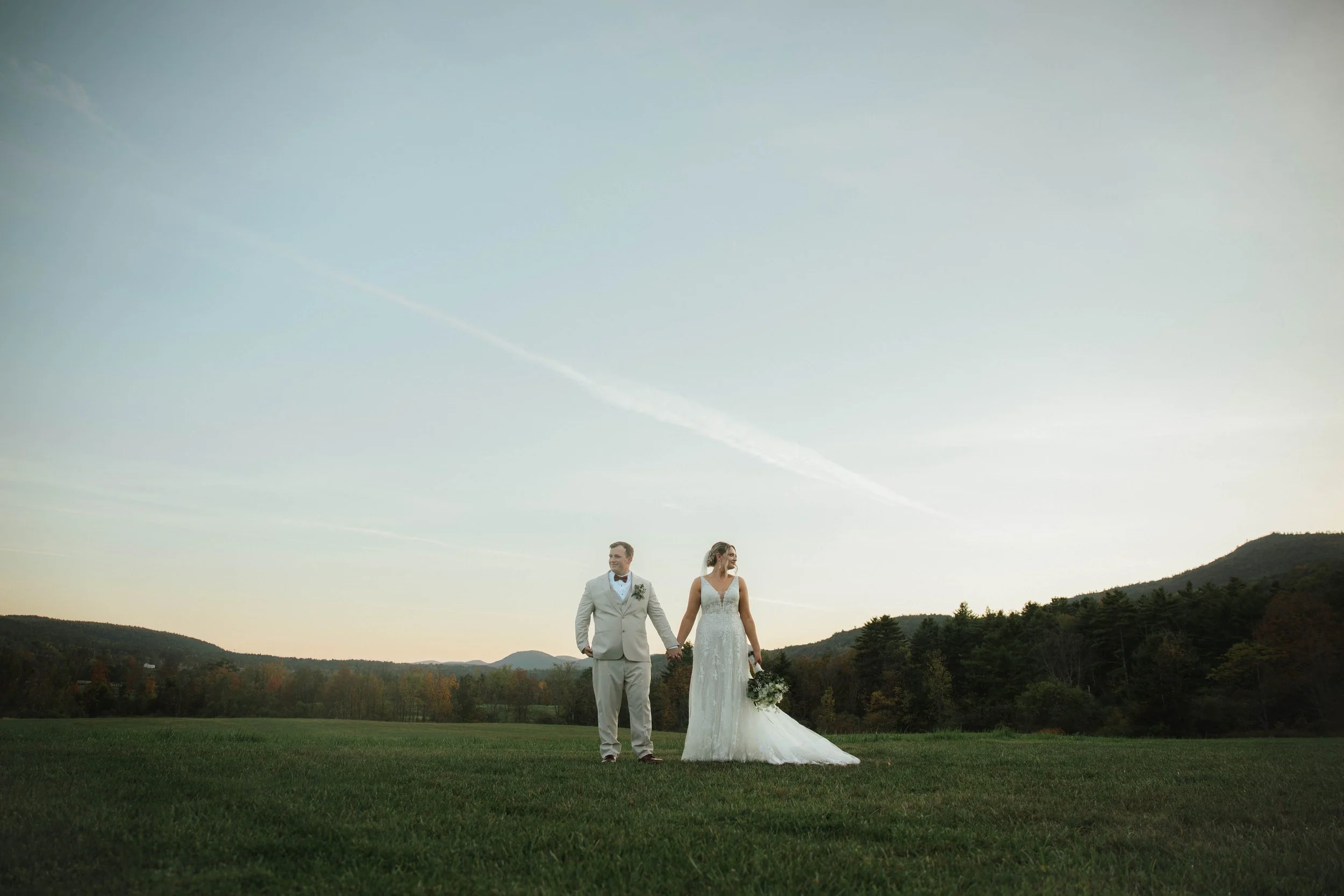 A bride and groom holding hands in a field at sunset, with trees and hills in the background.