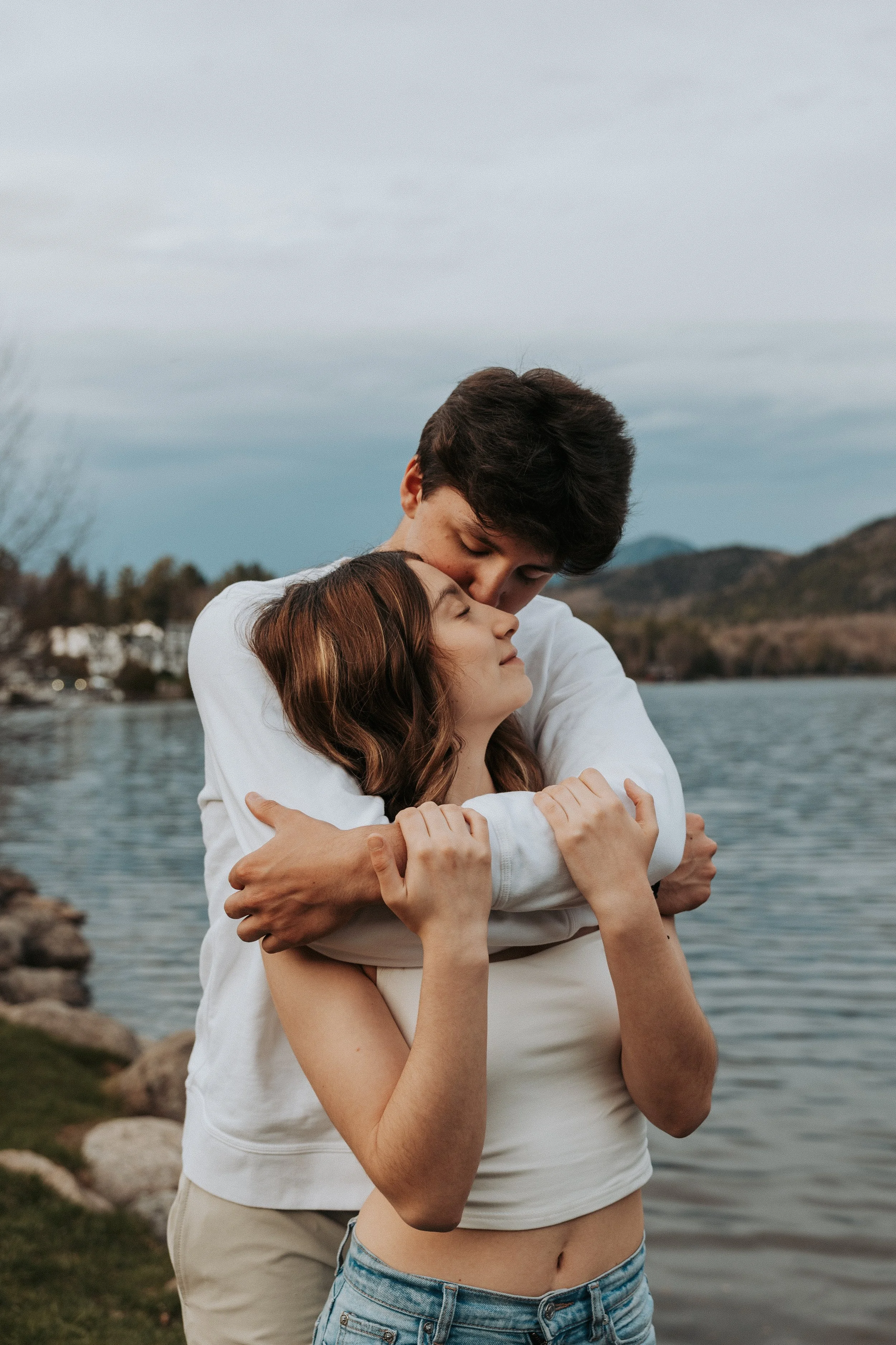 A young couple embracing by a lake on a cloudy day, with mountains in the background.