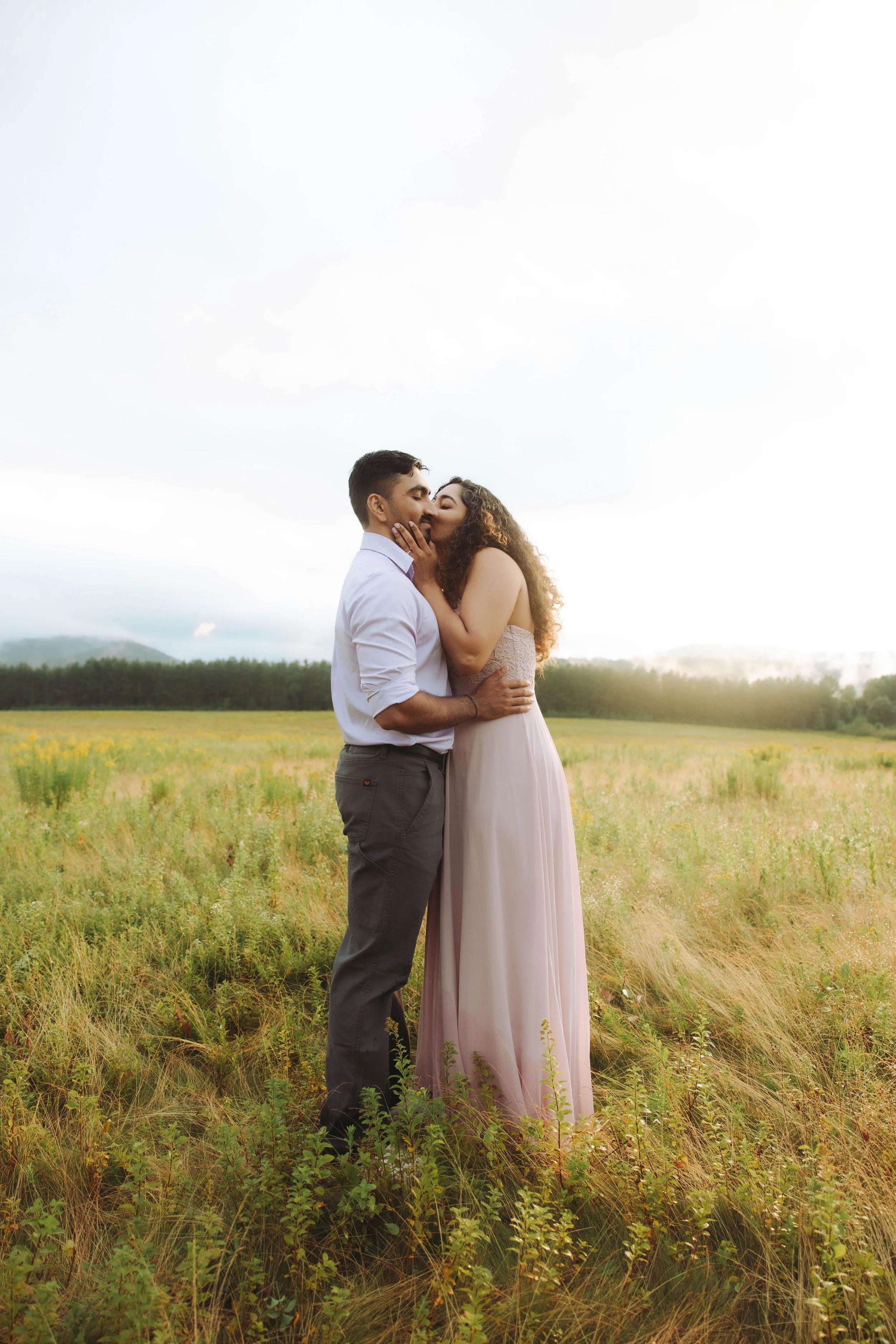 A couple sharing a kiss in a field during sunset, with a cloudy sky and distant trees in the background.