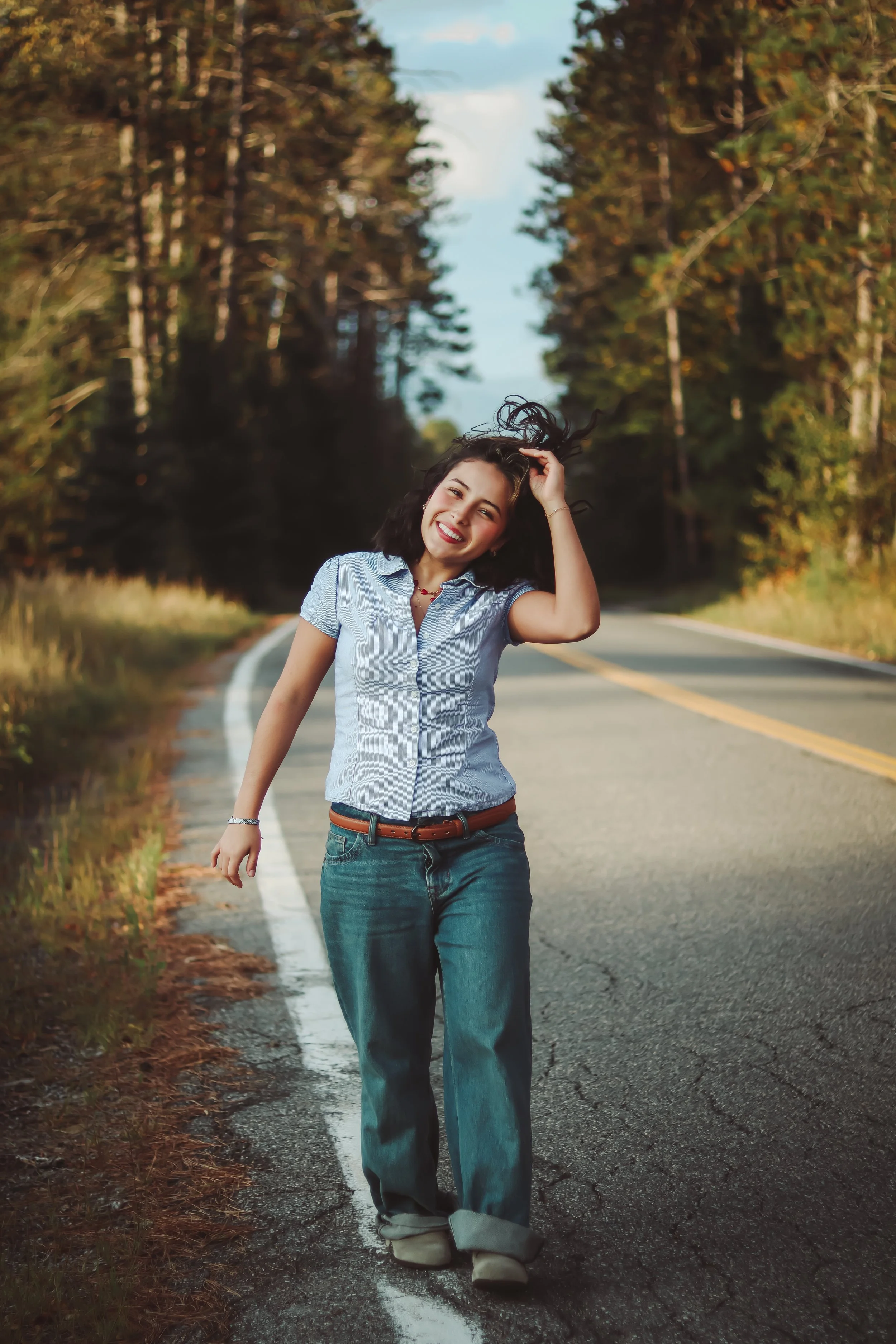 A young woman with black hair, wearing a blue button-up shirt and jeans, smiling and walking along a rural road with trees on both sides during daytime.
