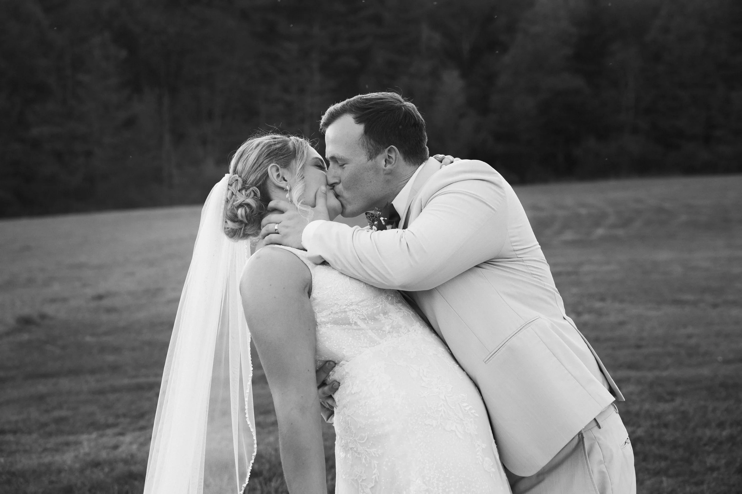 A black and white photograph of a bride and groom sharing a kiss outdoors, with a grassy field and trees in the background.