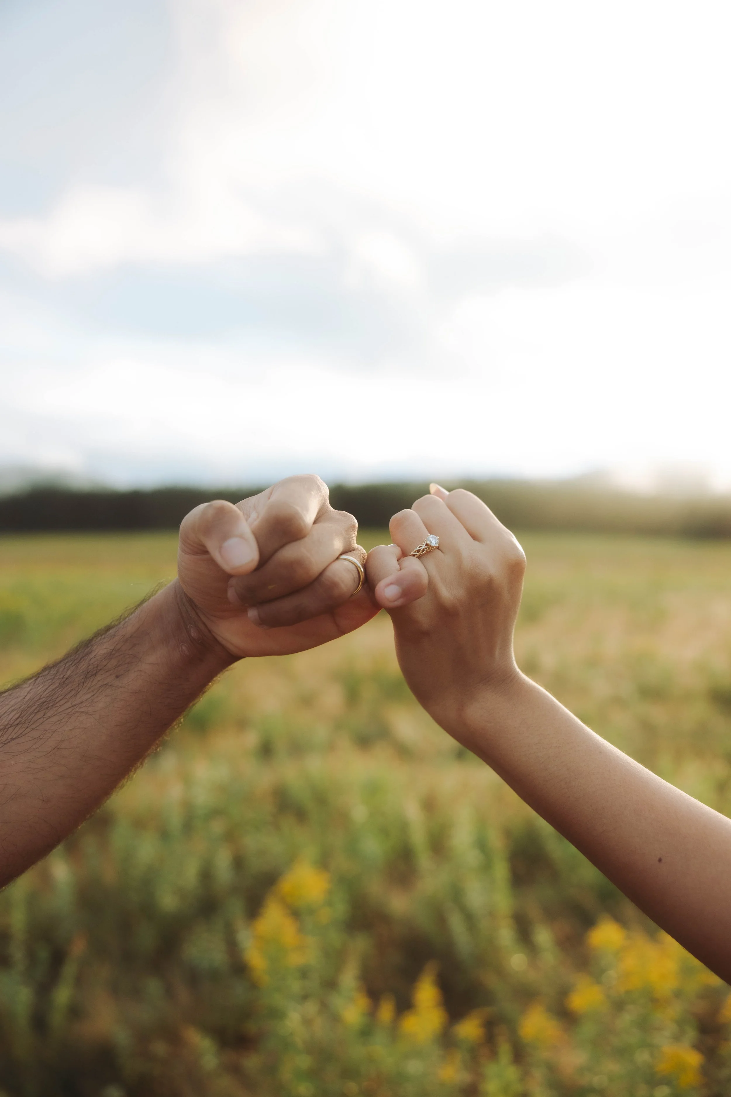 Two hands with wedding rings holding each other in an open field under a partly cloudy sky.