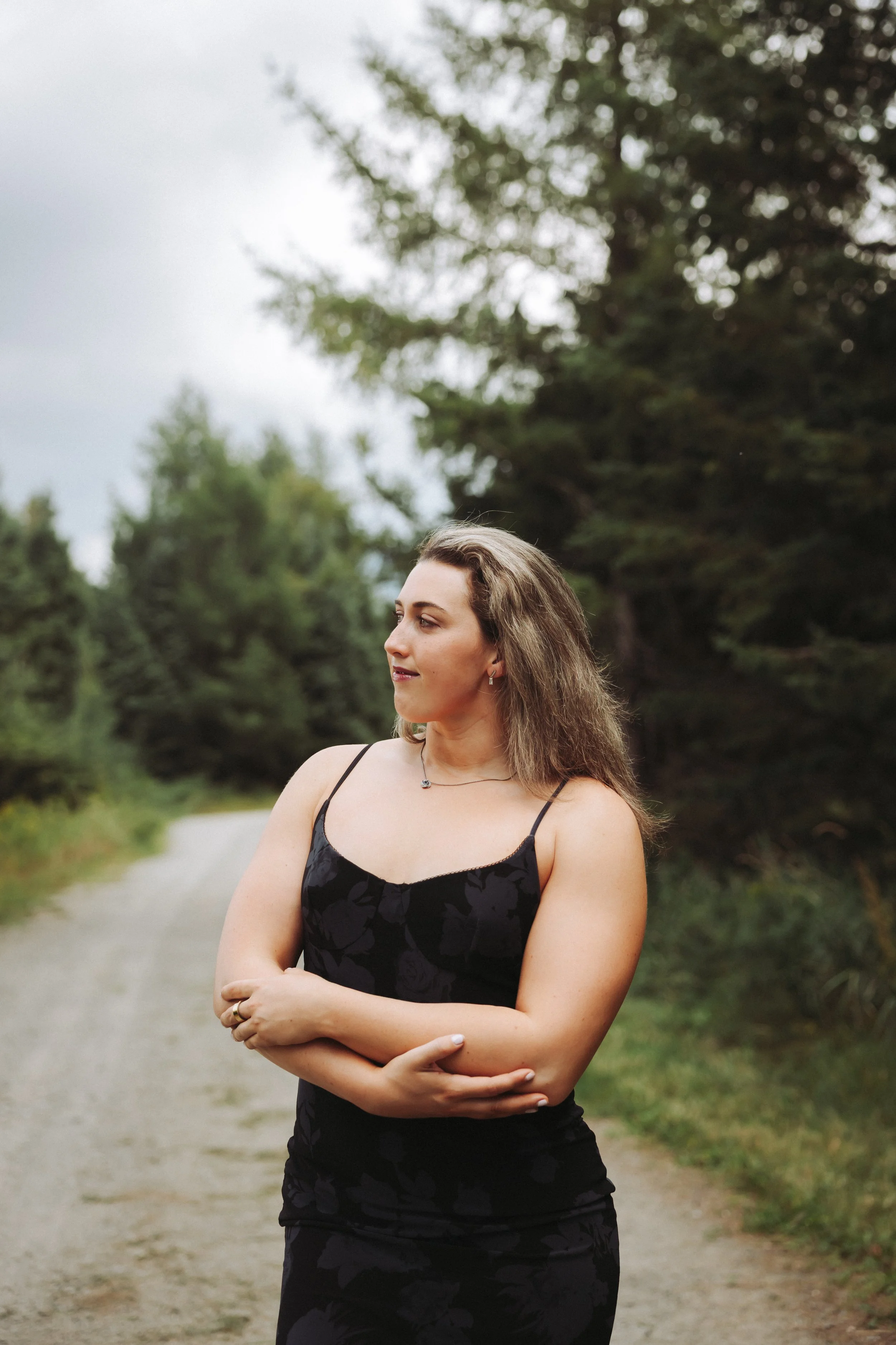 A woman in a black dress stands outdoors on a gravel path with trees in the background, looking to the side.