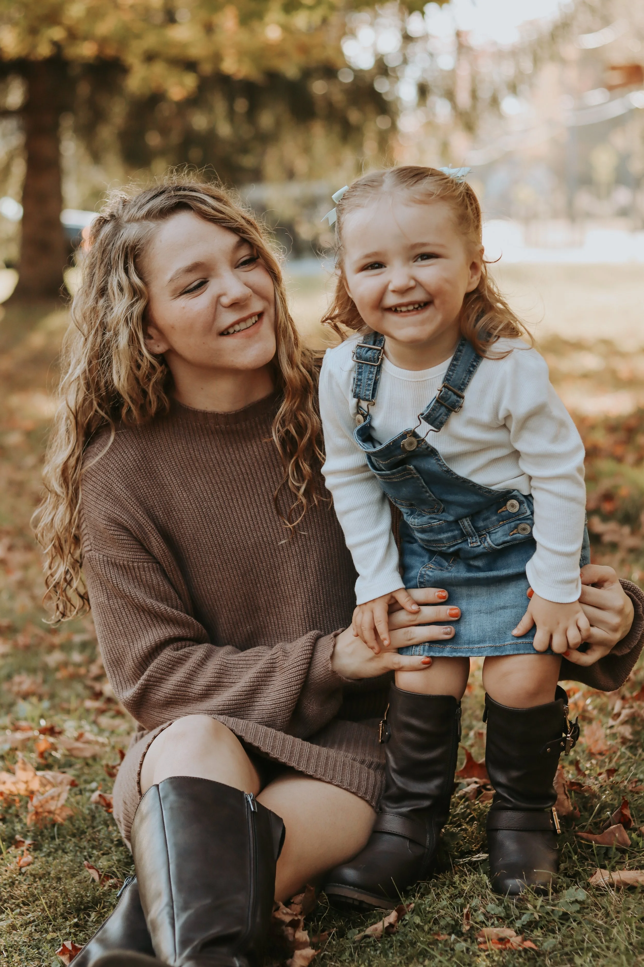 A woman and a young girl smiling outdoors in fall, the woman sitting on the ground with the girl standing in front of her, holding her legs, surrounded by autumn leaves.