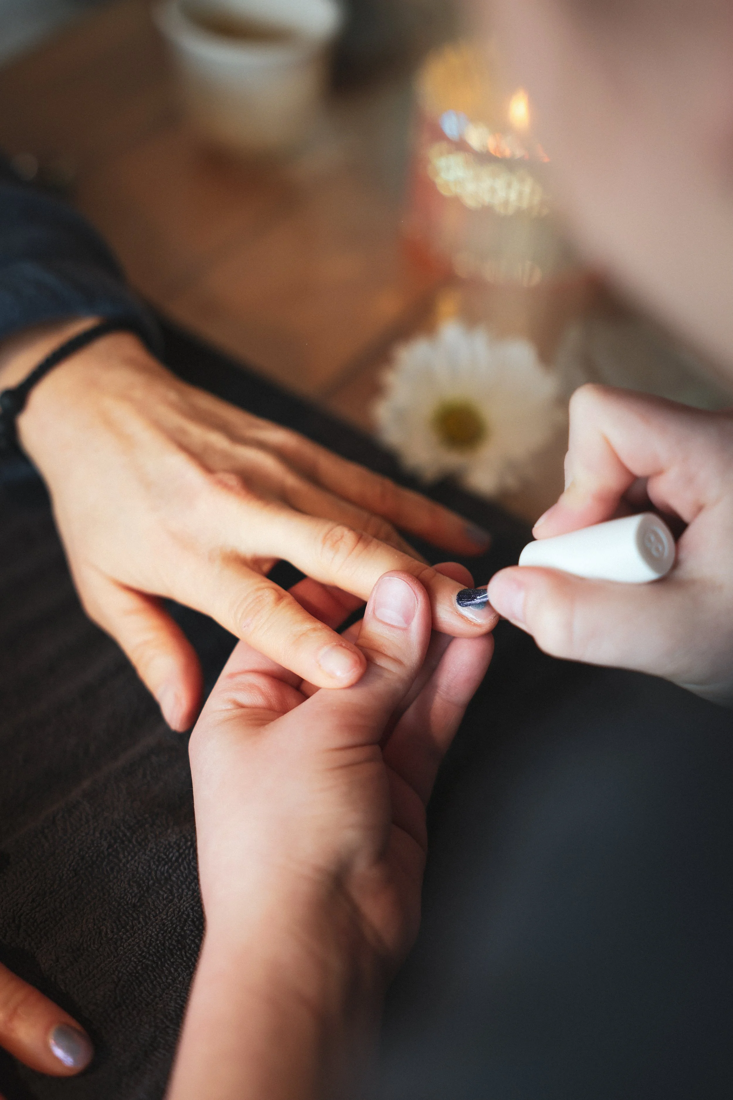 Close-up of a person painting a child's fingernail with nail polish, with a daisy flower and blurred background visible.