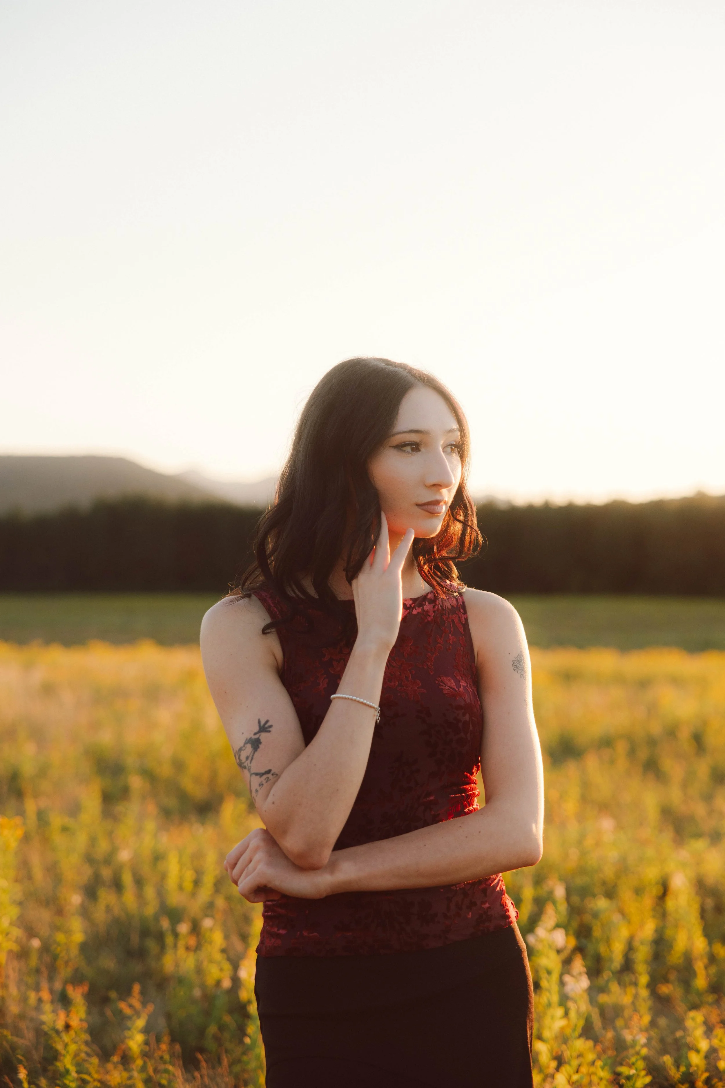 A young woman with dark, wavy hair stands in a field at sunset, wearing a sleeveless burgundy top with black patterns, looking thoughtfully to the side with her hand near her face.