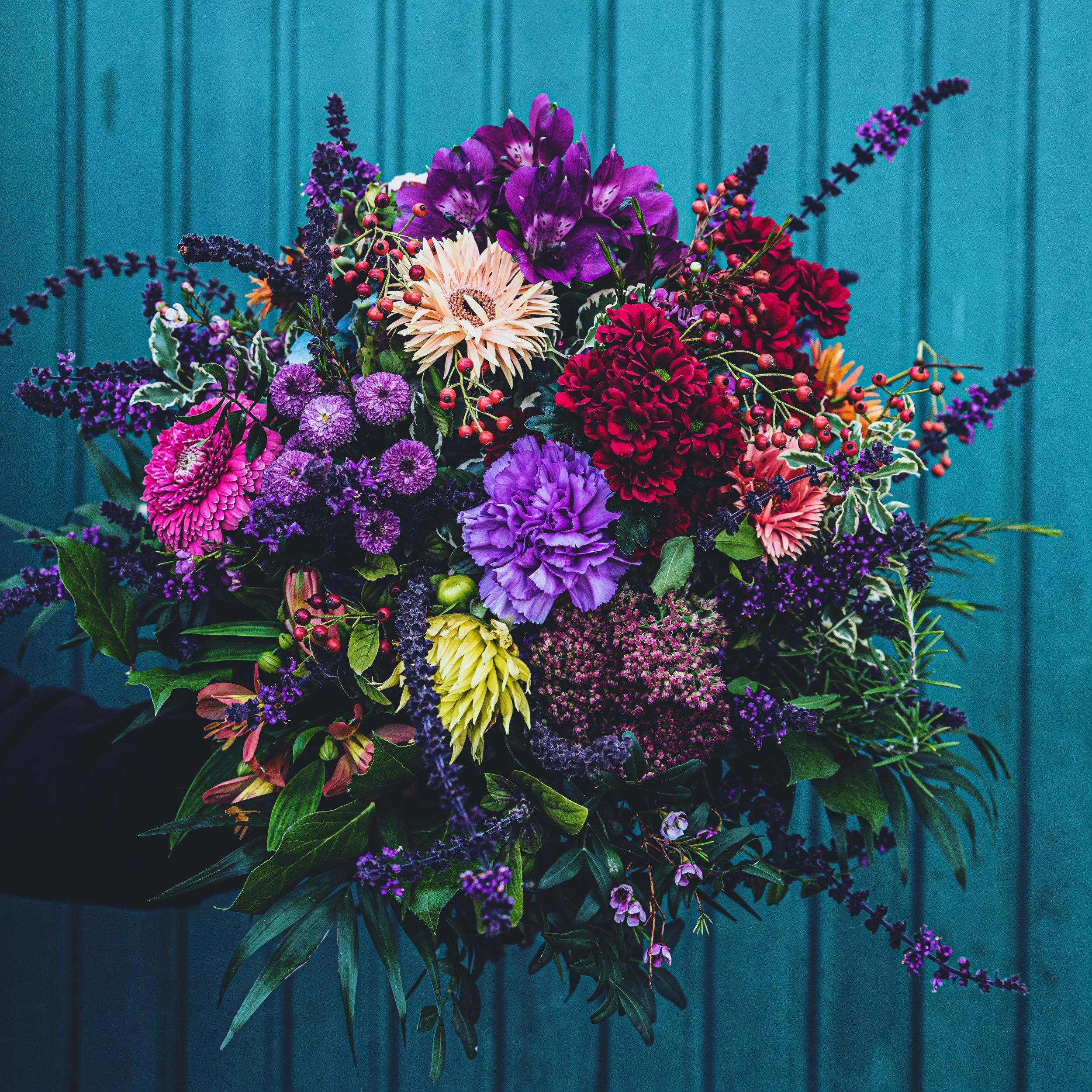 Bunter Blumenstrauß mit violetten, pinken, roten und gelben Blumen vor einer blauen Wand.