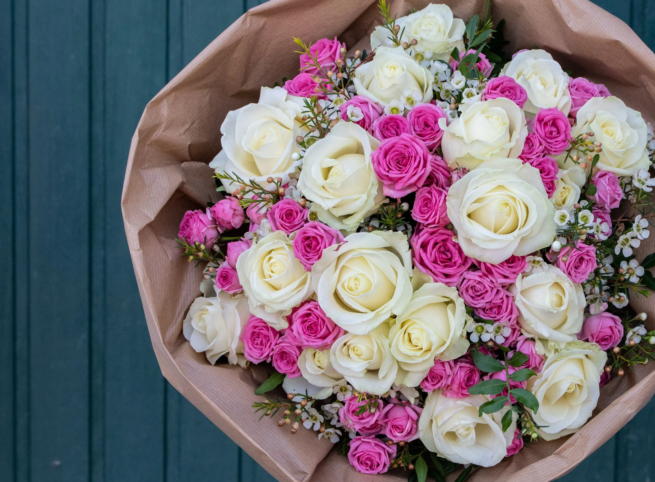 Ein bunter Blumenstrauß aus weißen und pinken Rosen, umgeben von kleinen weißen Blüten und grünem Blattwerk, in brauner Packpapier eingepackt auf einer blauen Holzfläche.