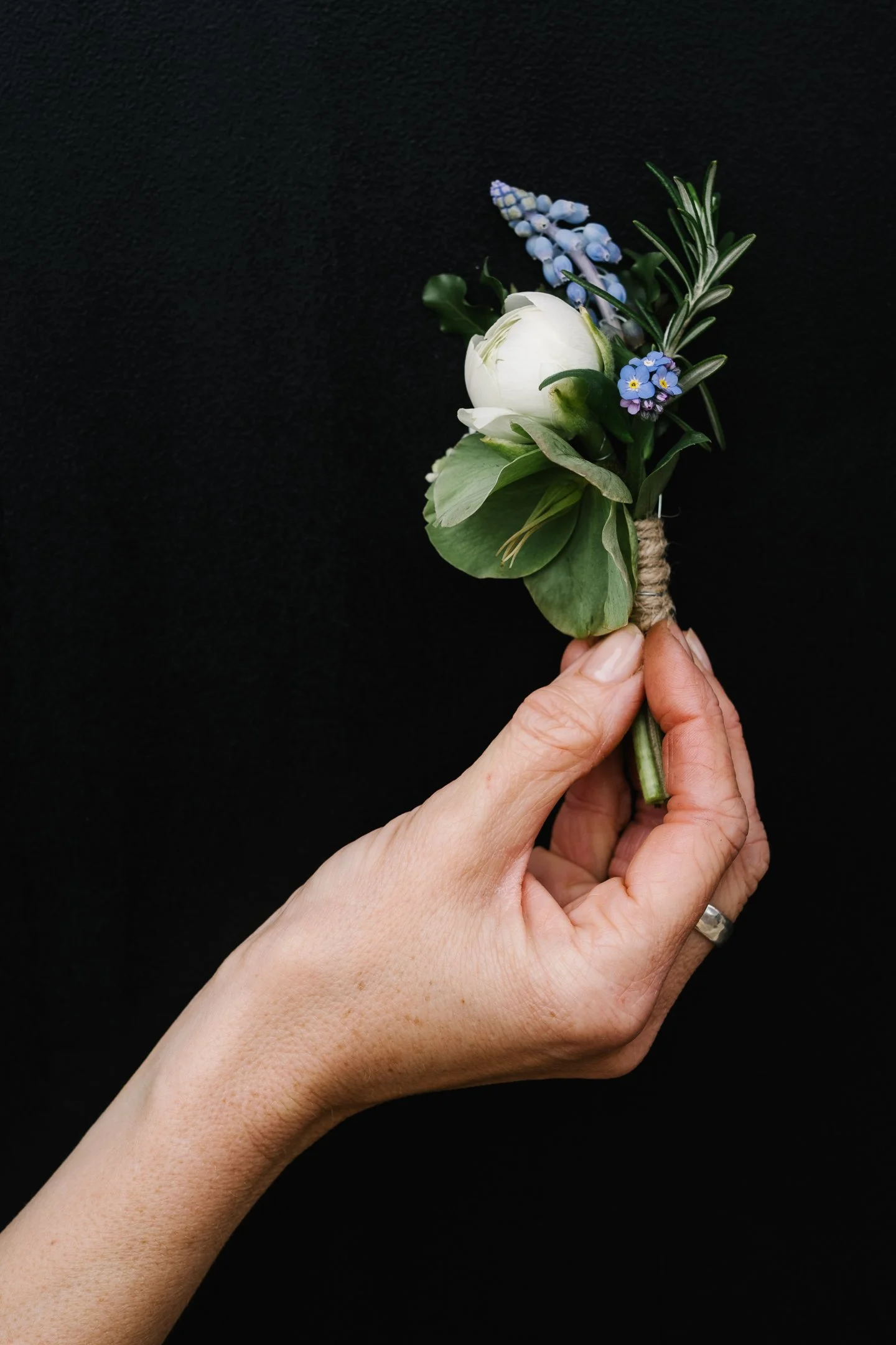 Hand holding a small floral boutonniere with a white flower, blue grape hyacinths, forget-me-nots, rosemary, and greenery against a black background