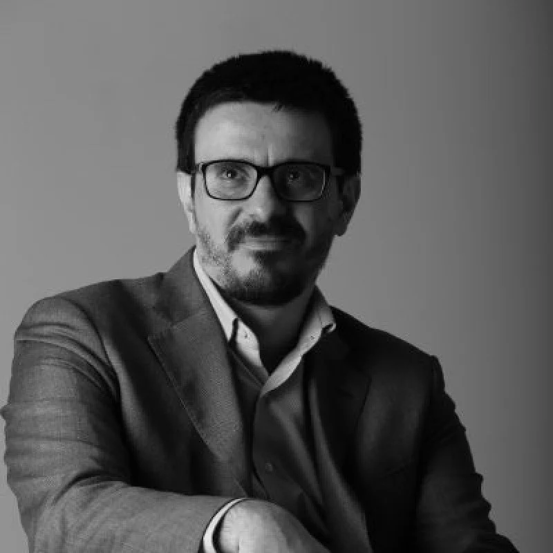 A black-and-white portrait of a man wearing glasses and a suit, with short dark hair and facial hair, against a plain background.