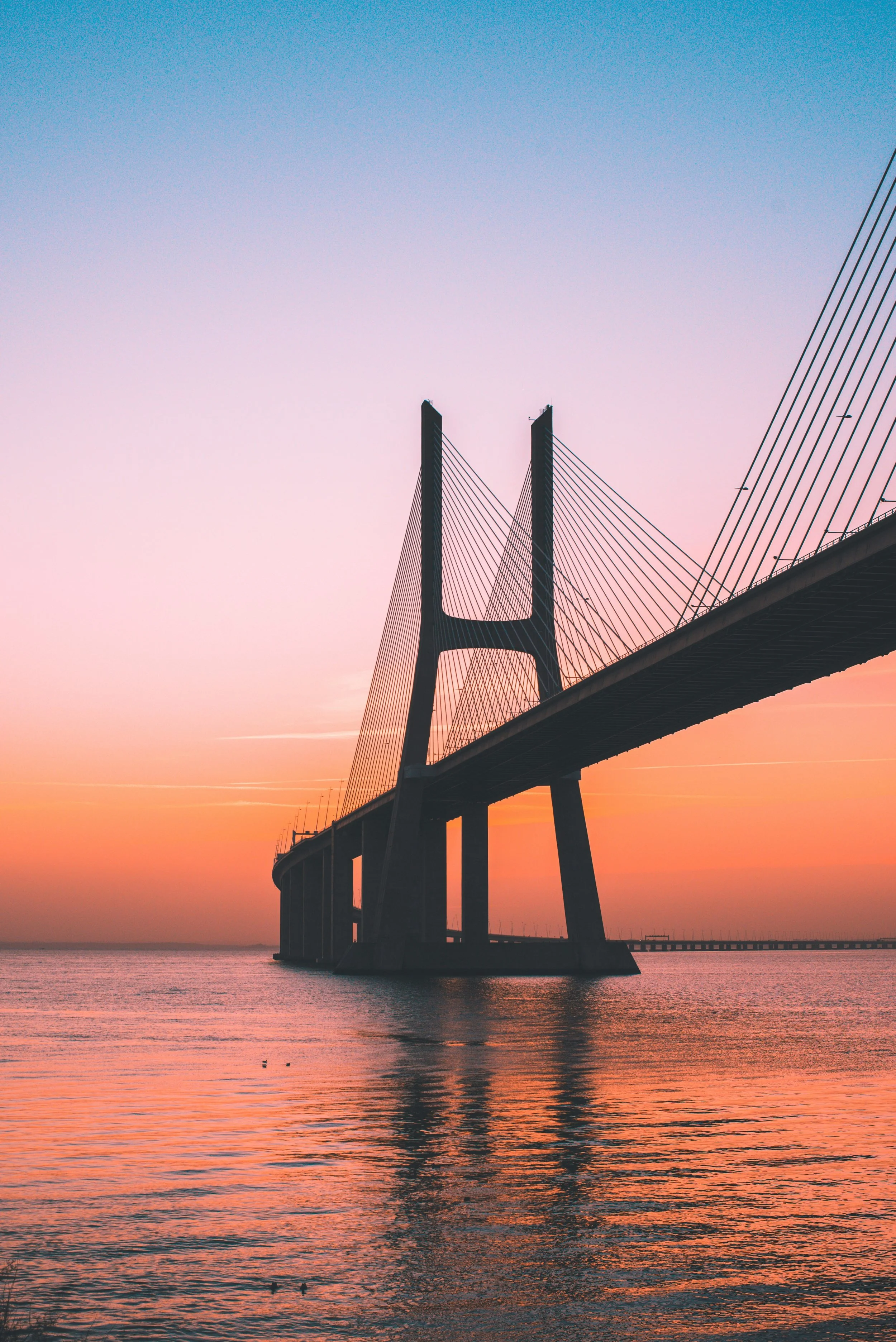 A suspension bridge extending over water at sunset with a gradient sky transitioning from pink to blue.