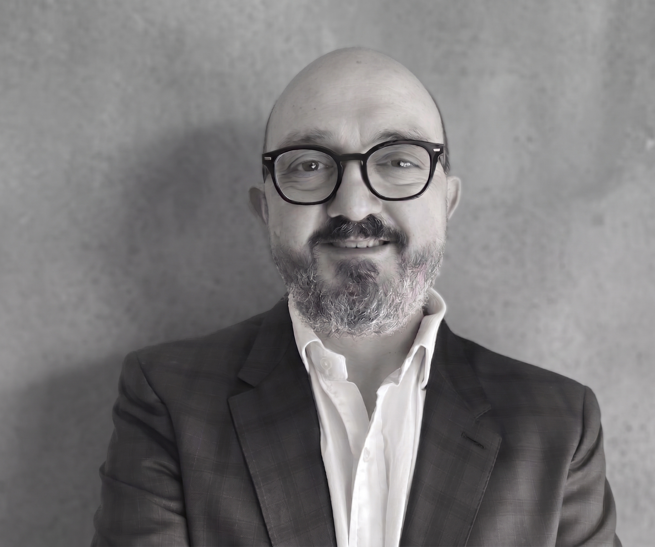 A close-up of a man with a bald head and grey beard, wearing black glasses, a white shirt, and a dark checkered blazer, standing against a grey textured background.