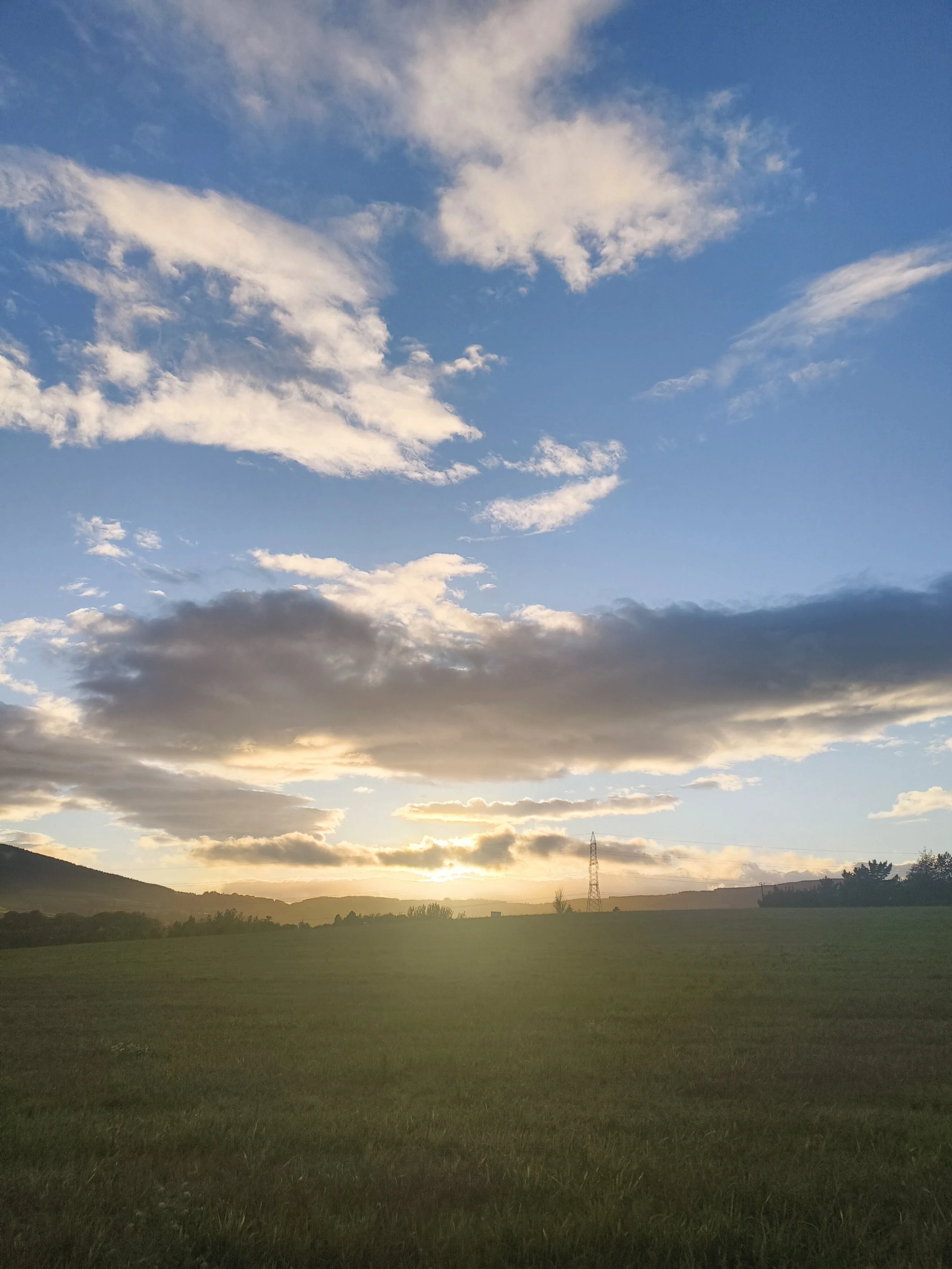 A landscape scene during sunrise or sunset with green fields in the foreground, hills in the distance, and a partly cloudy sky with the sun near the horizon.