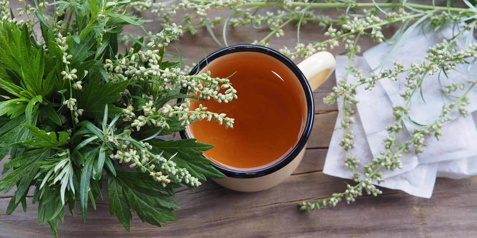 A cup of herbal tea surrounded by fresh mint and possibly basil on a wooden surface.