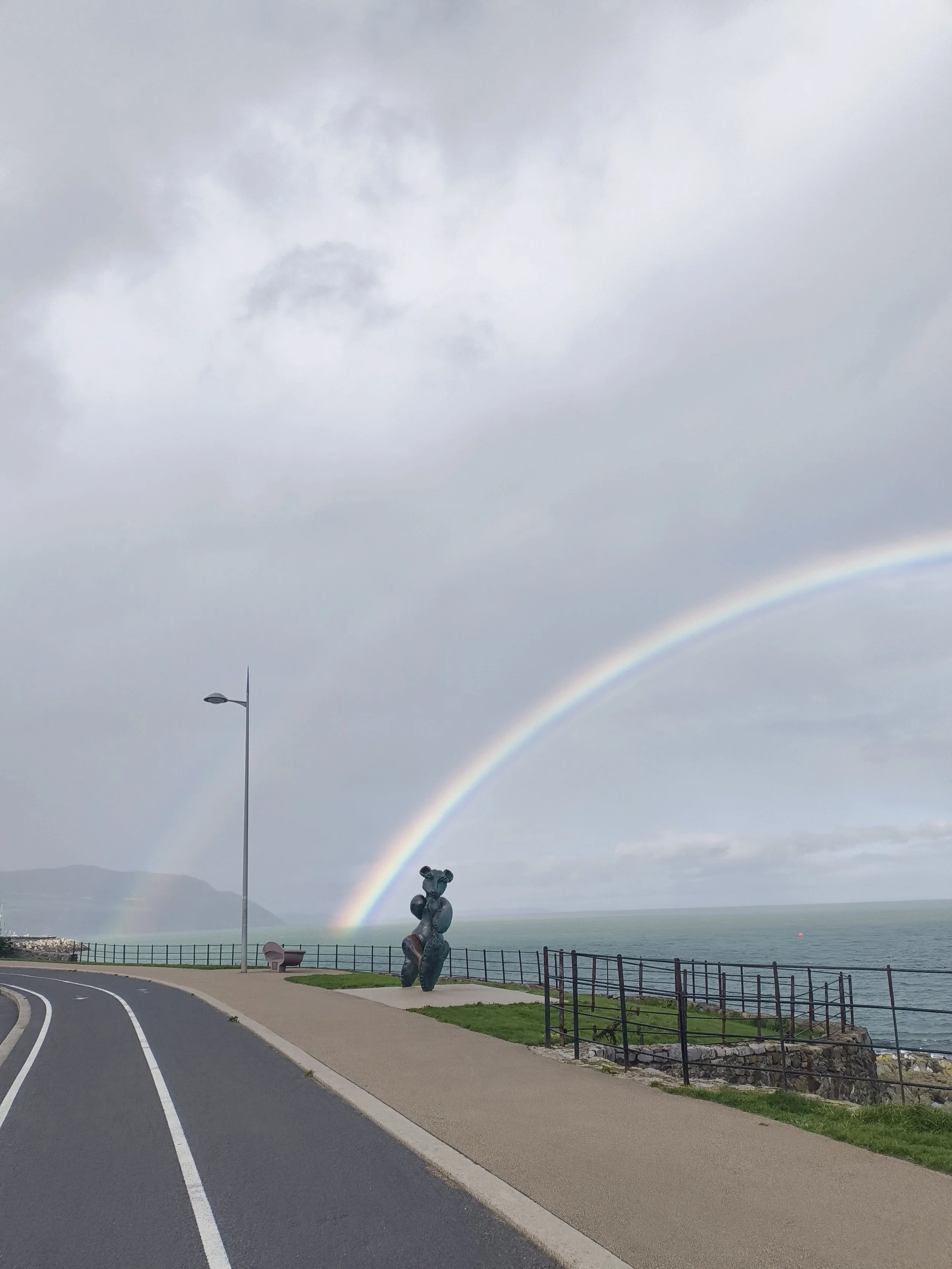A scenic lakeside view with a rainbow arching across a cloudy sky, a sculpture resembling a bear or animal on the pathway, a fence along the water, and a lamppost beside the road.