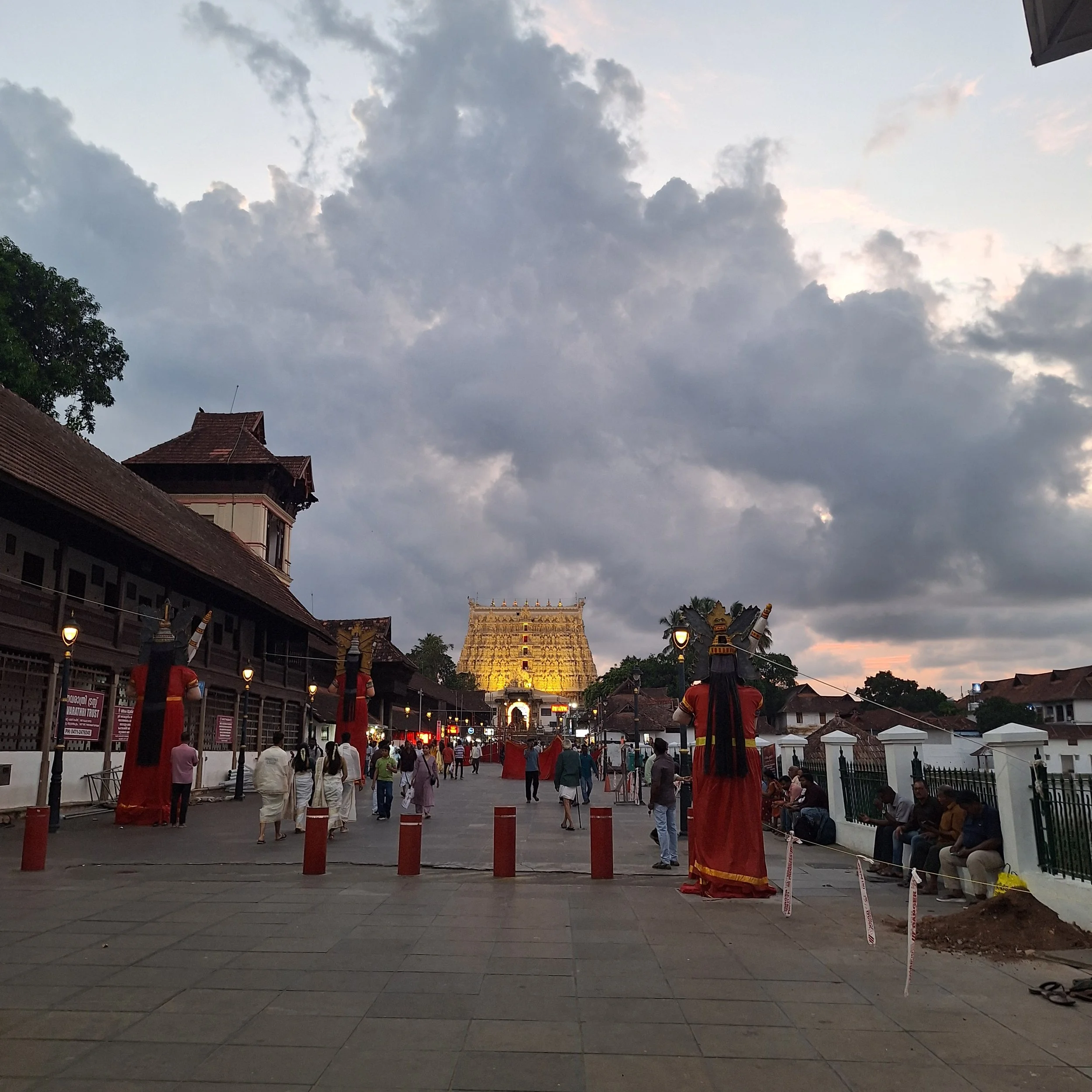 A busy street scene near a Hindu temple with a large, lit-up gopuram (temple tower) in the background. People are walking, some in traditional attire, with decorations and red lanterns along the street, and a cloudy sky overhead.