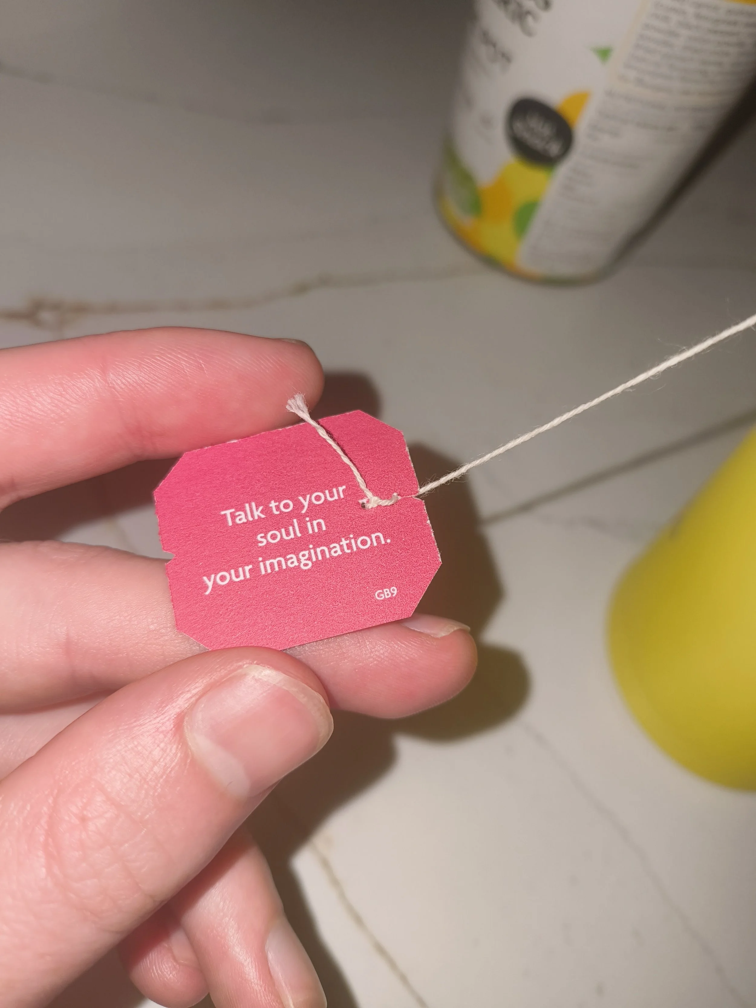 Close-up of a person's fingers holding a pink paper tag with a white string attached. The tag has white text that says: 'Talk to your soul in your imagination.' In the background, partial view of a yellow object and a cylindrical container with a col