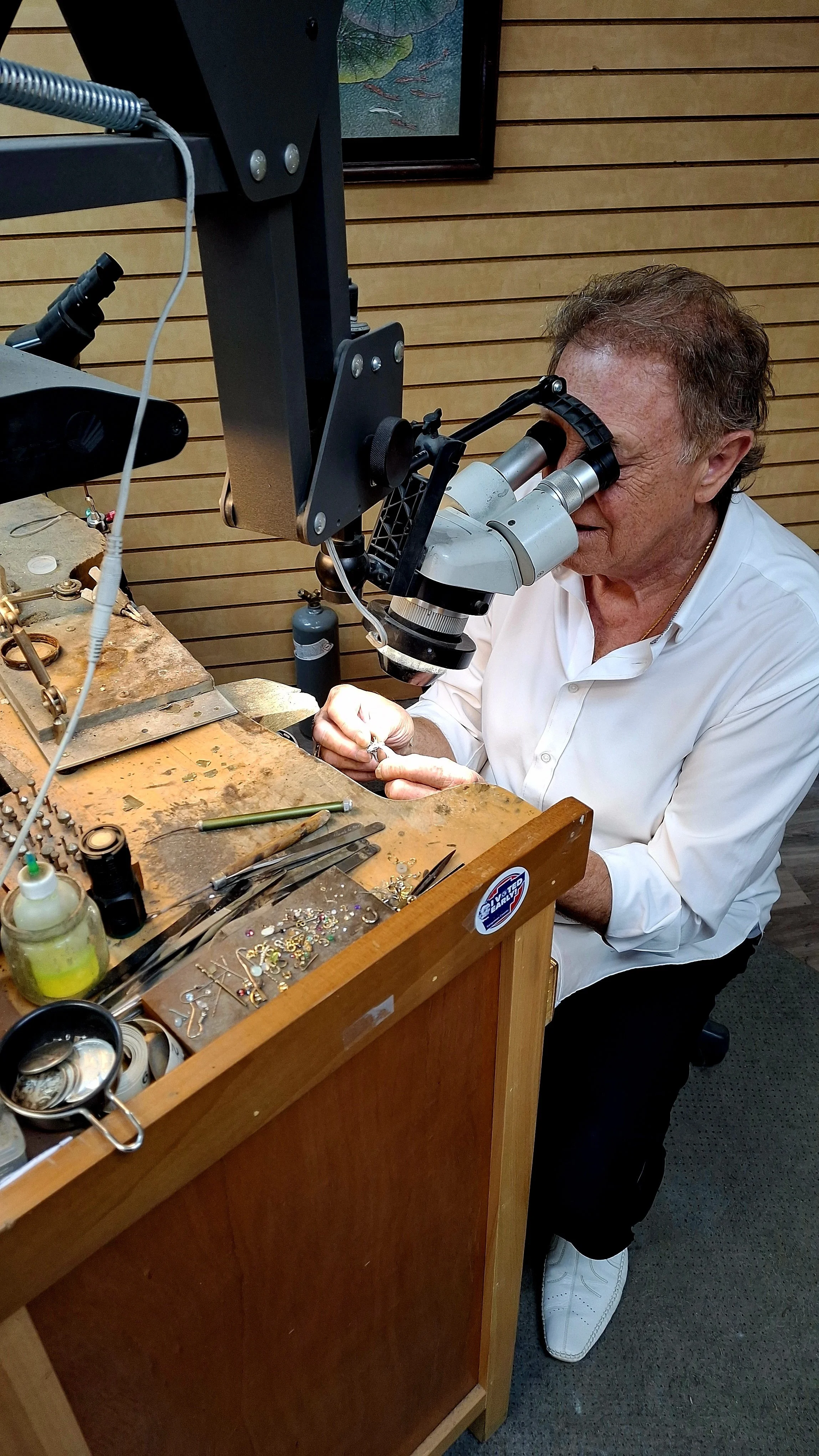 A man using a microscope to examine jewelry or small objects at a workbench filled with jewelry-making tools and supplies.