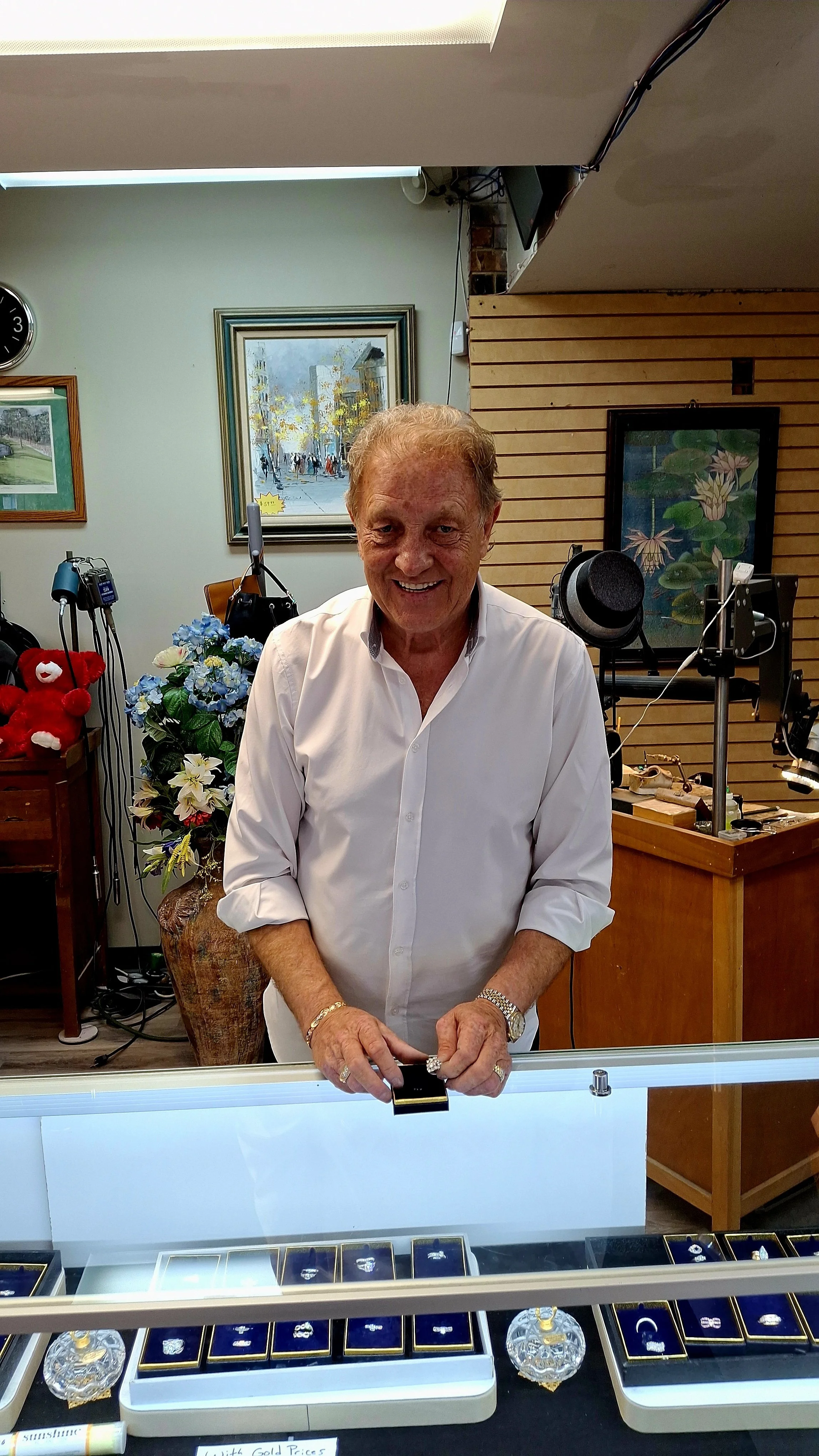 A smiling elderly man in a white shirt standing behind a jewelry display case with rings and rings in a jewelry store.